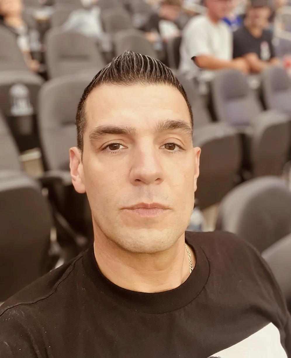 A close-up selfie of a man with dark hair slicked back, wearing a black shirt, sitting in an auditorium with gray seats. Other people are visible in the background.