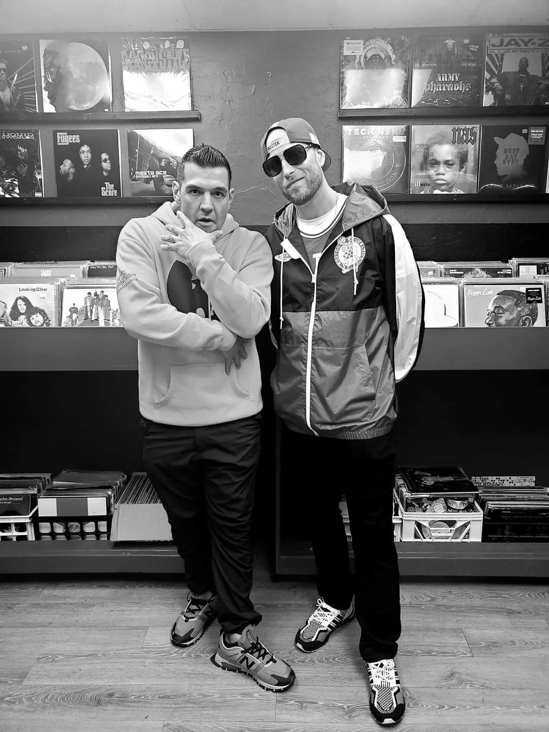 Two men standing in front of a display of vinyl records in a store. One man is wearing a hoodie, sneakers, and has glasses on. The other man is wearing a jacket, sunglasses, and a cap.
