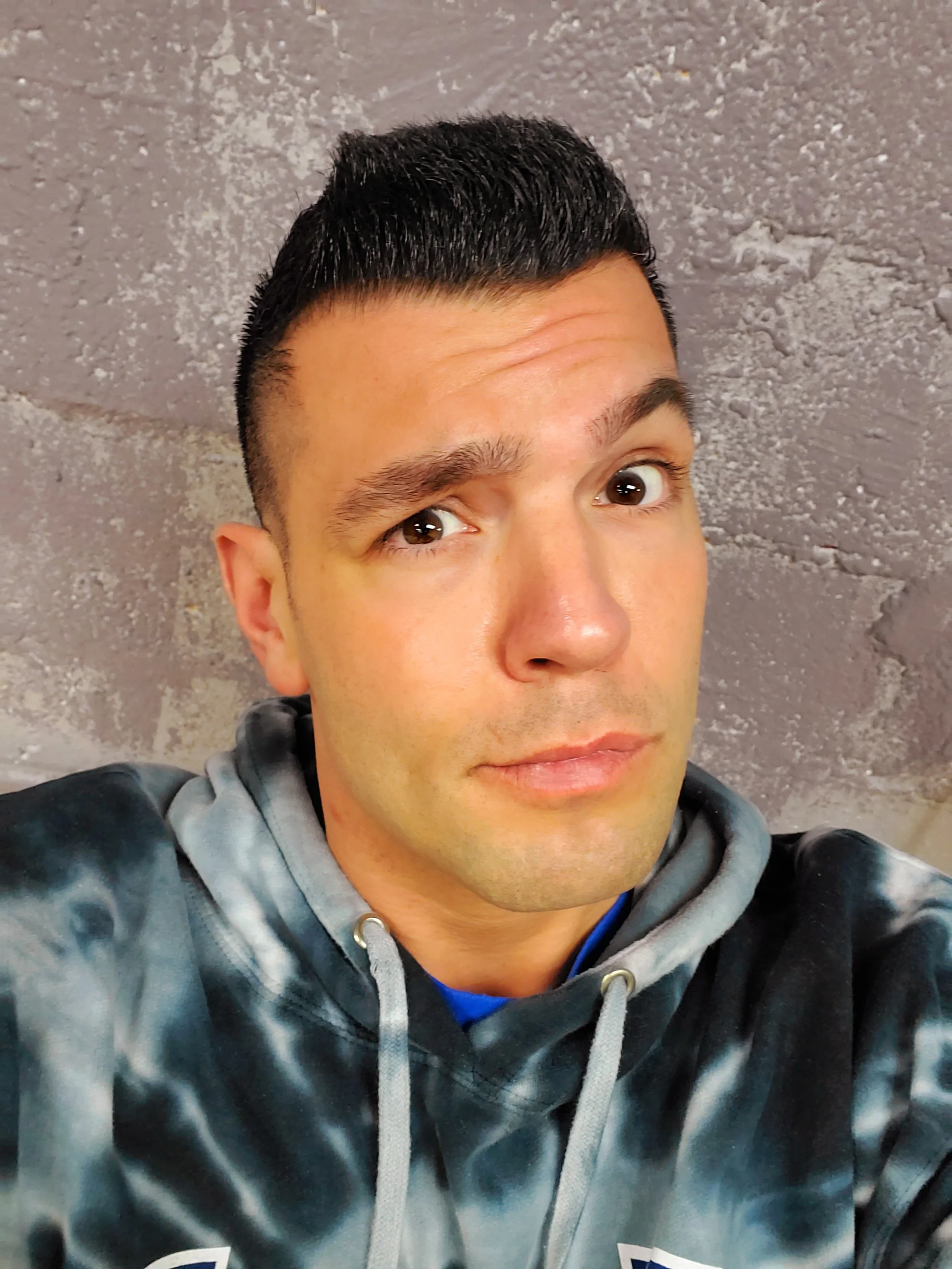 A young man with short black hair and brown eyes wearing a black and gray tie-dye hoodie taking a selfie against a gray brick wall.