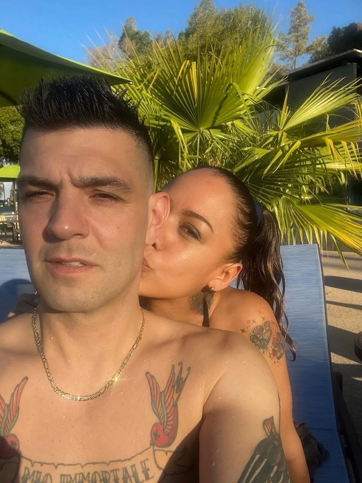 A shirtless man with tattoos on his chest taking a selfie outdoors with a woman kissing his cheek, with a background of palm trees and sunny sky.