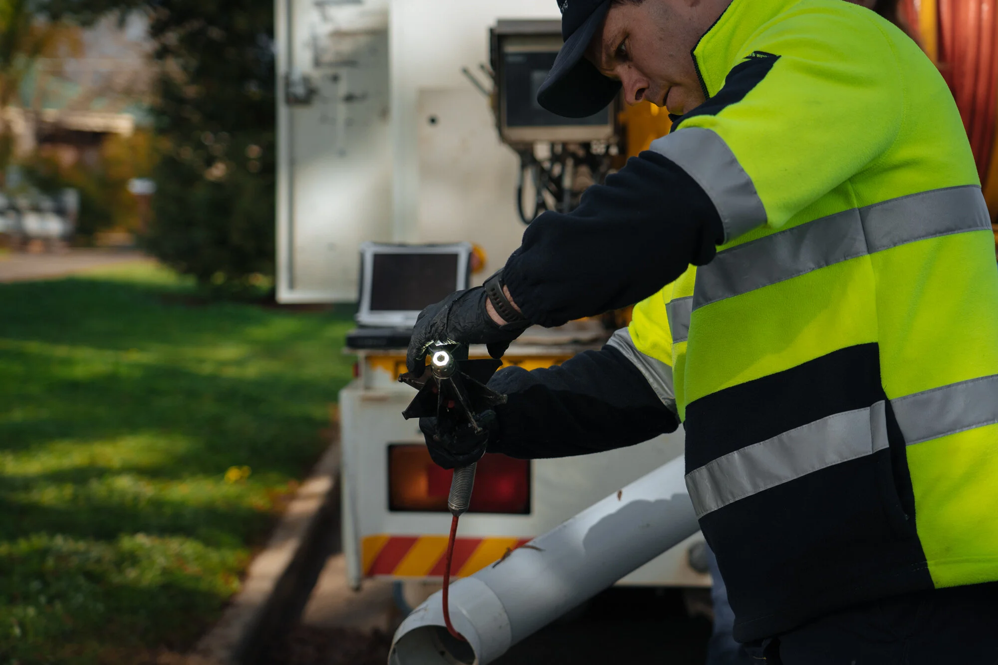 A utility worker in a high-visibility yellow safety vest is using a tool to repair or install equipment on a utility pole or service truck in an outdoor setting.