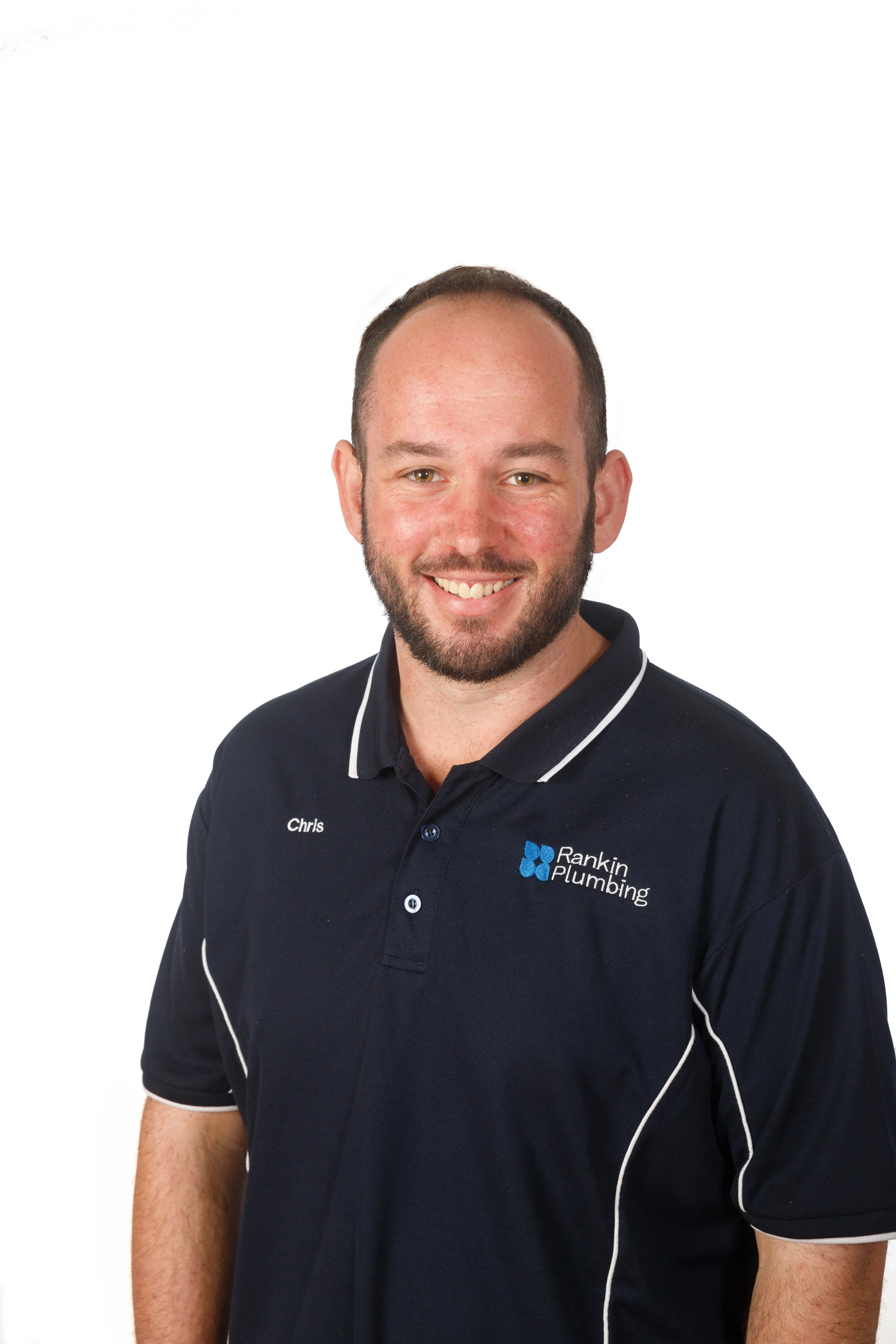 A man wearing a navy polo shirt with white piping and a logo that reads 'Rankin Plumbing' on the left chest; he has a beard, short hair, and is smiling.