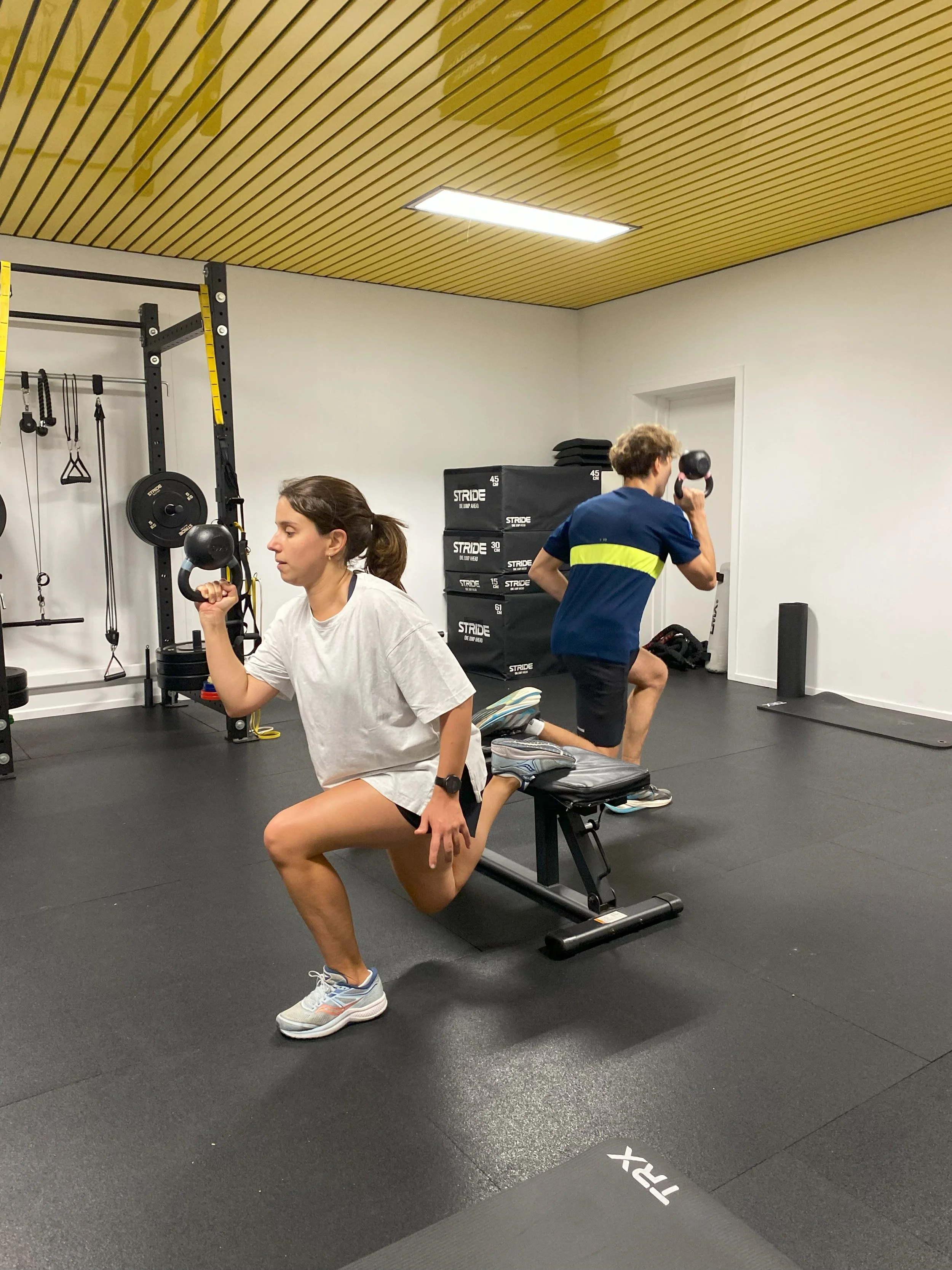 Twee mensen in een fitnessruimte die met kettlebells aan het trainen zijn. Vrouw zit op een oefenbank en houdt een kettlebell, man staat achter haar met een kettlebell bij het oor.