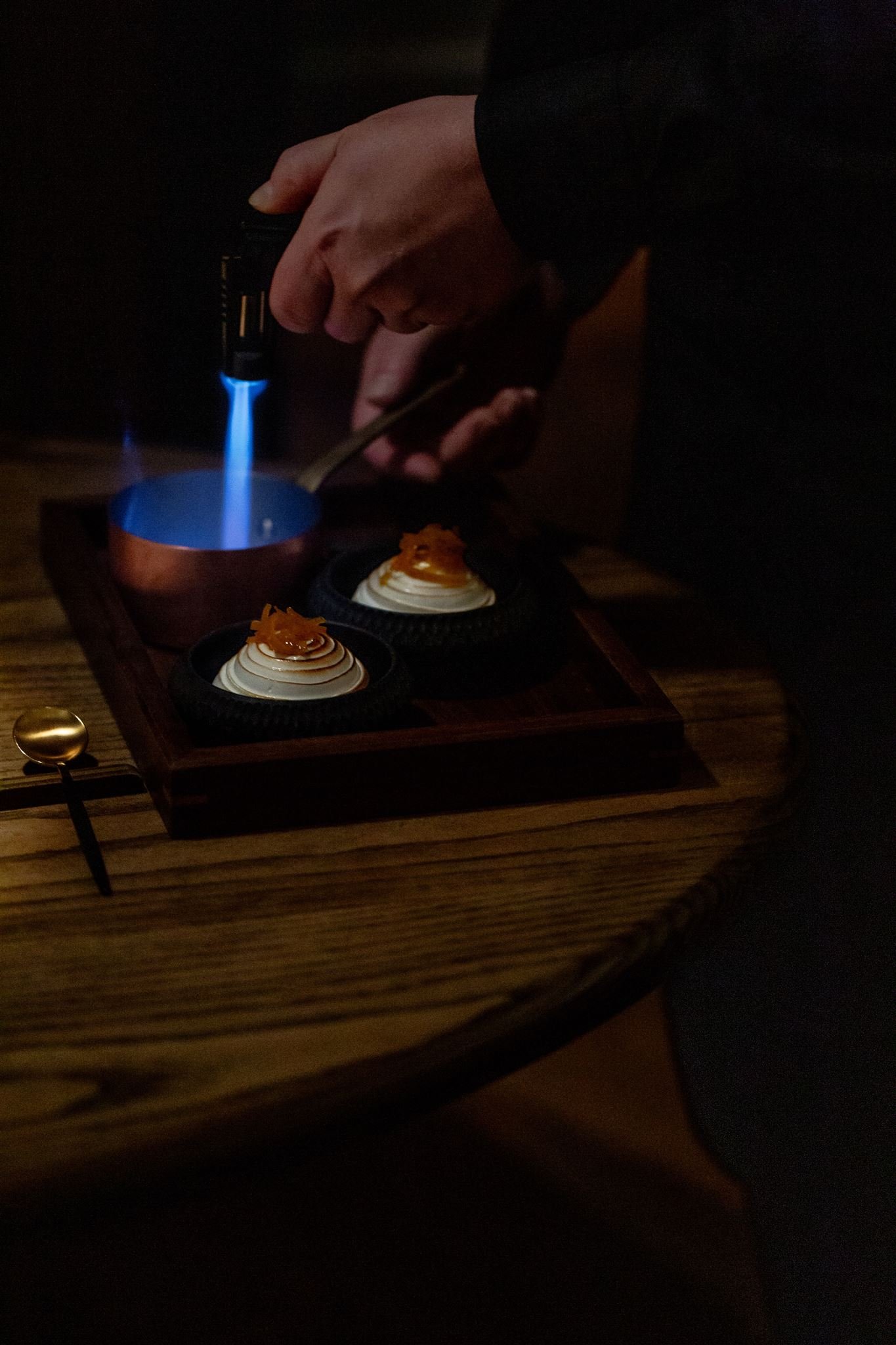 Two plated desserts on a wooden tray while a cook torches a sauce