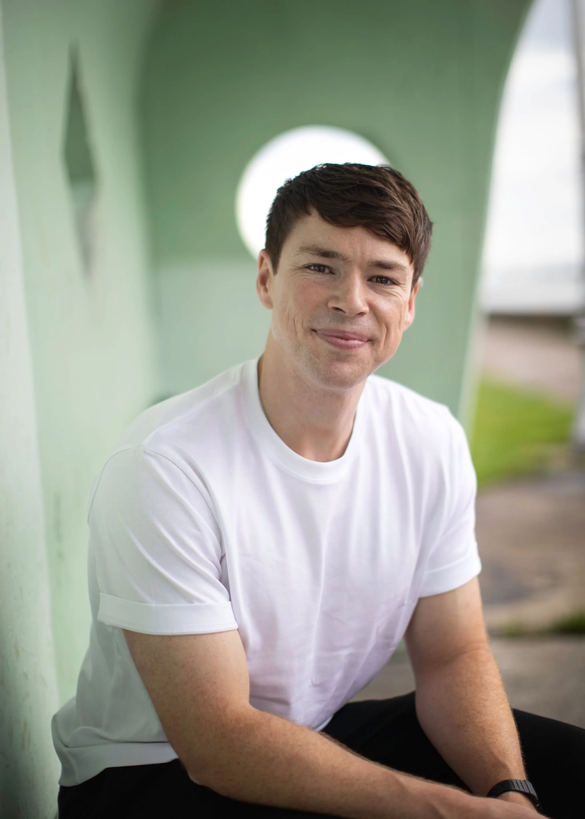 A young man with dark hair and a white T-shirt, smiling and sitting against a green wall with circular cutouts.
