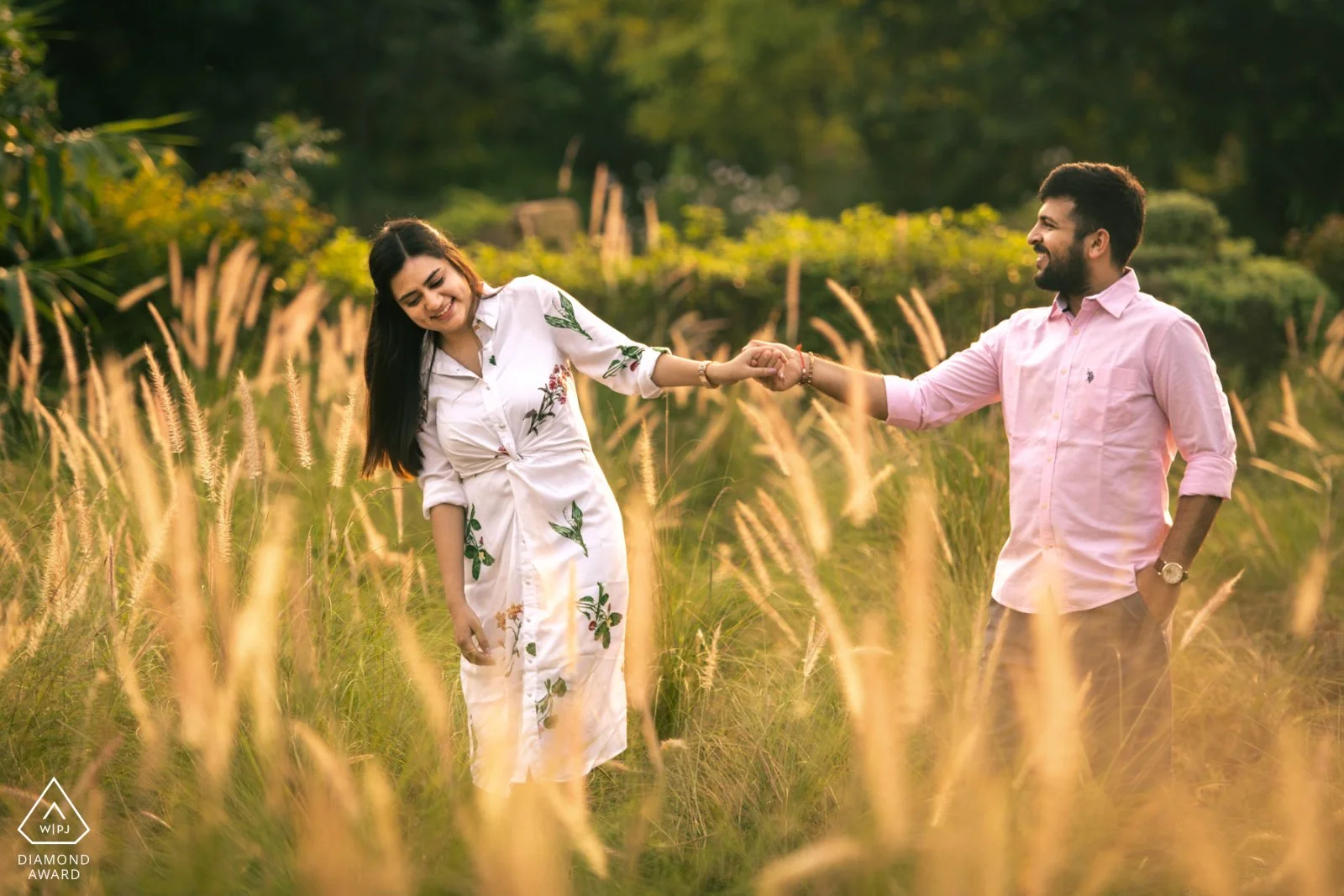 A young woman and man holding hands and smiling in a grassy field during sunset, with trees in the background.