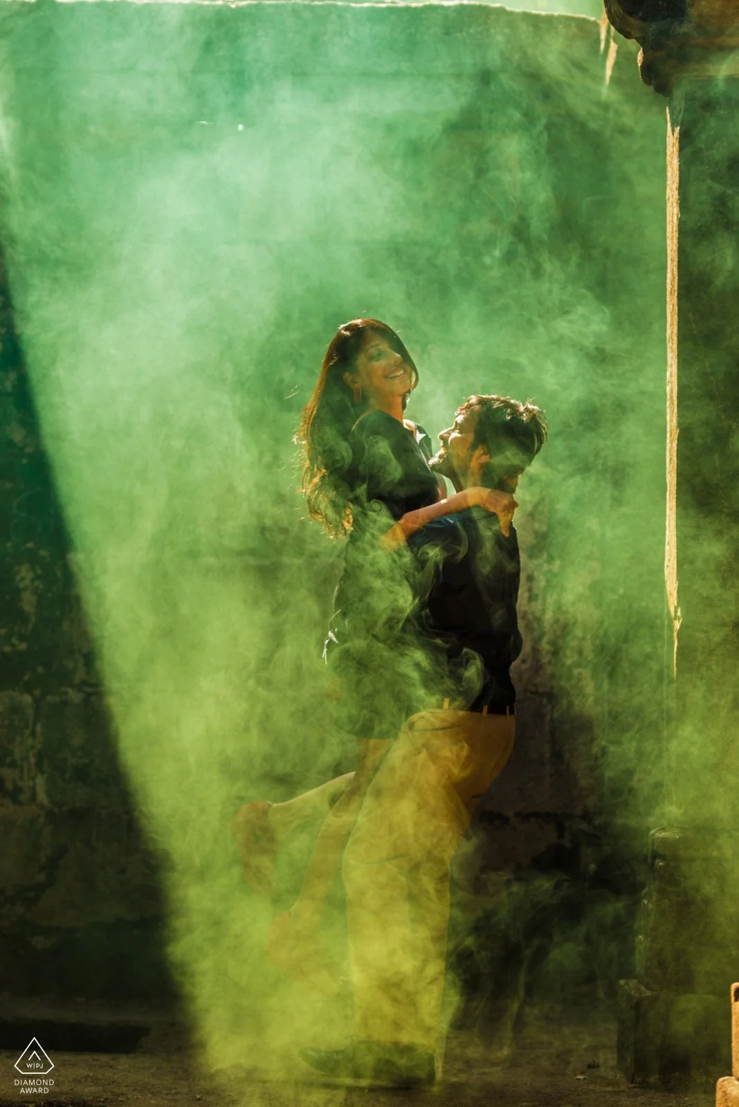 A man lifts a woman in the air, both smiling and looking happy, with green smoke or fog surrounding them in an outdoor setting.