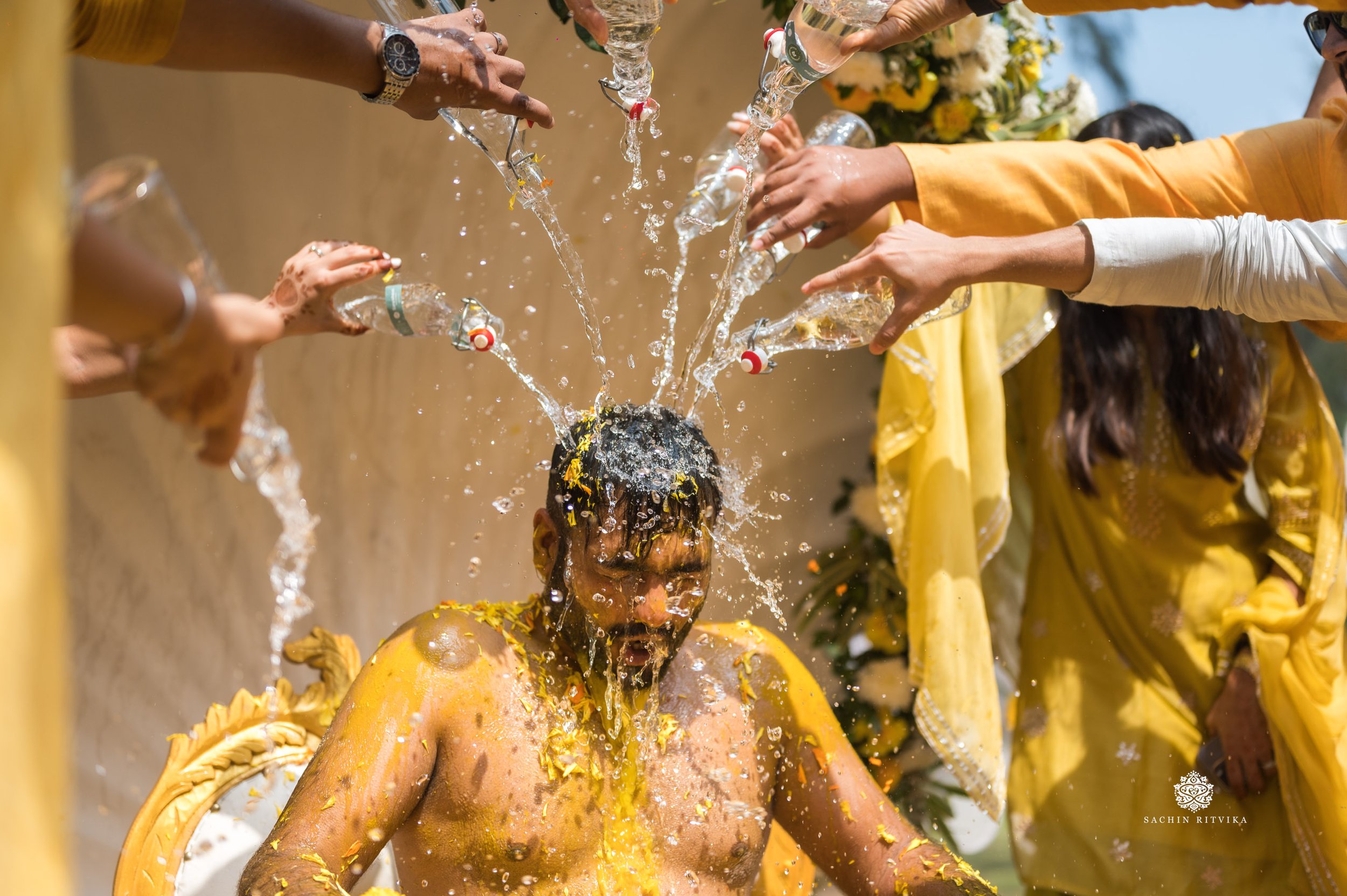 A traditional pre-wedding ceremony with people pouring water over a groom covered in yellow turmeric paste.