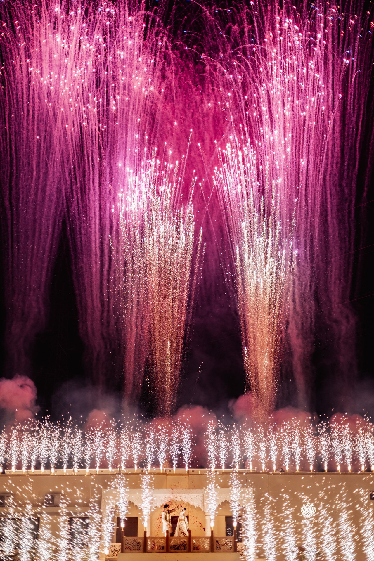 A couple embracing on a balcony beneath a large display of purple and pink fireworks at night.