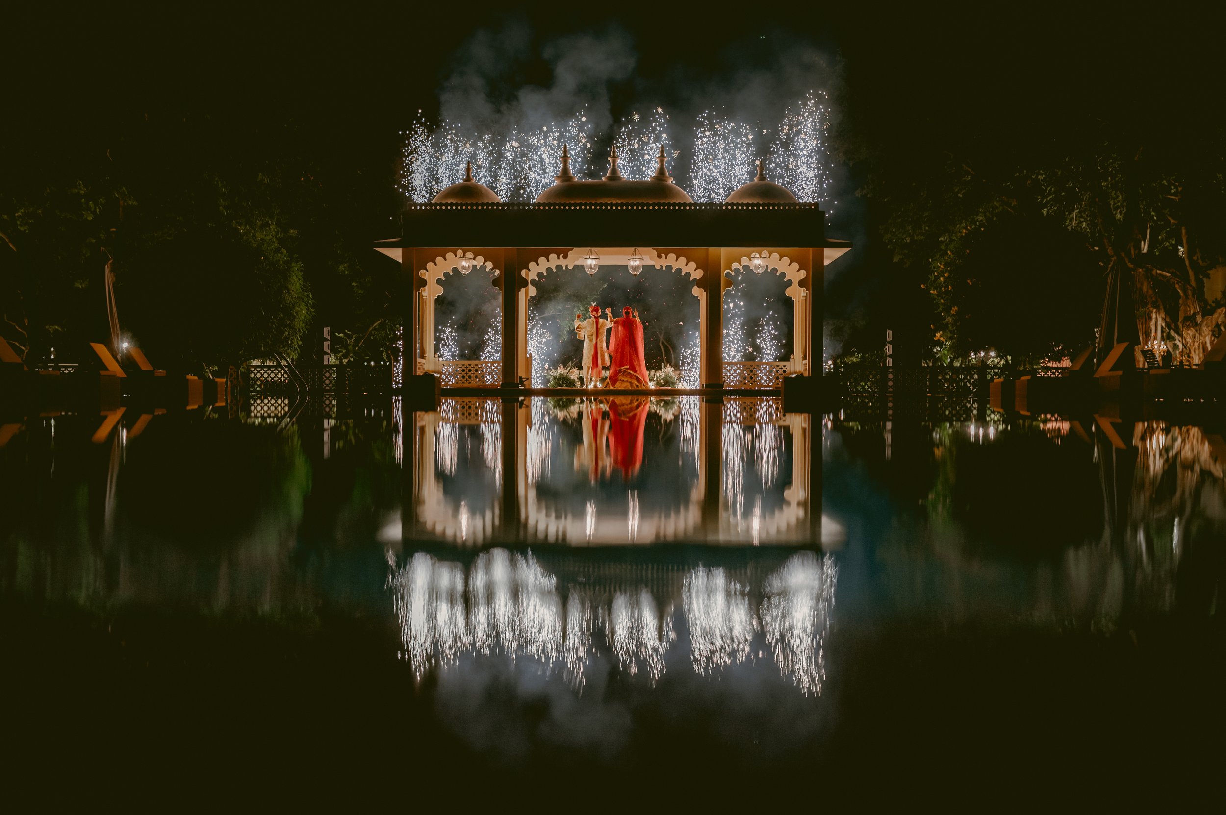 A couple in traditional attire stands under a lit pavilion with fireworks above and reflections in water below.