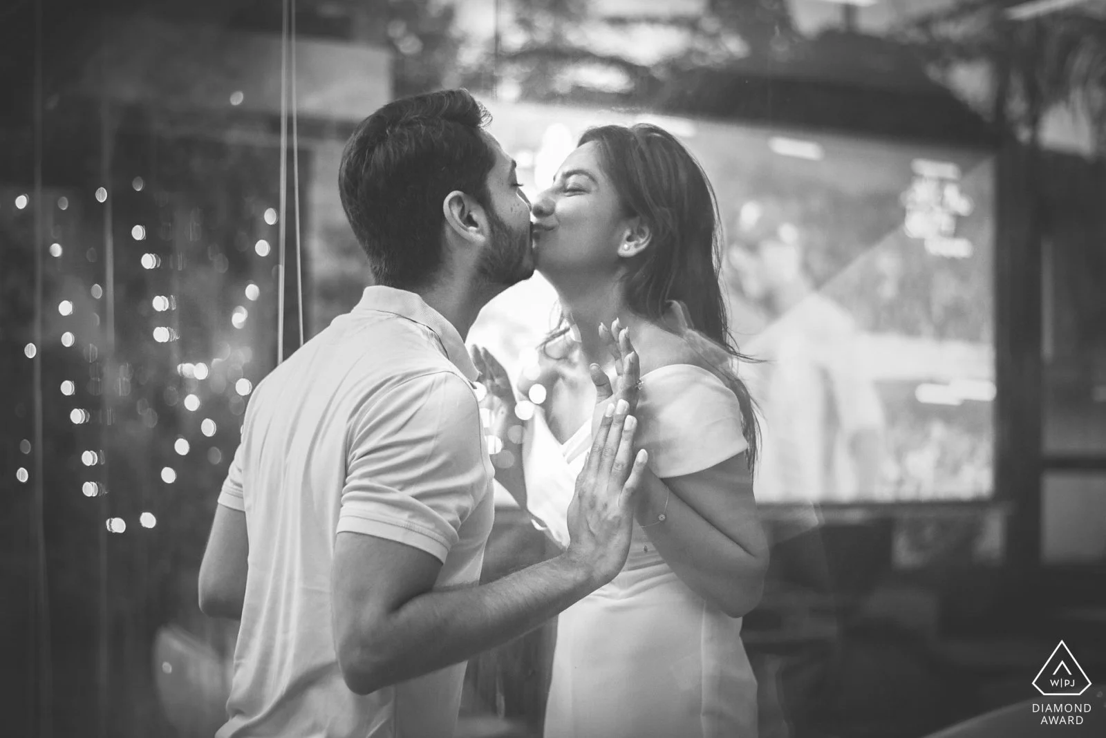 A black and white photo of a couple kissing through a glass window, with reflections and bokeh lights in the background.