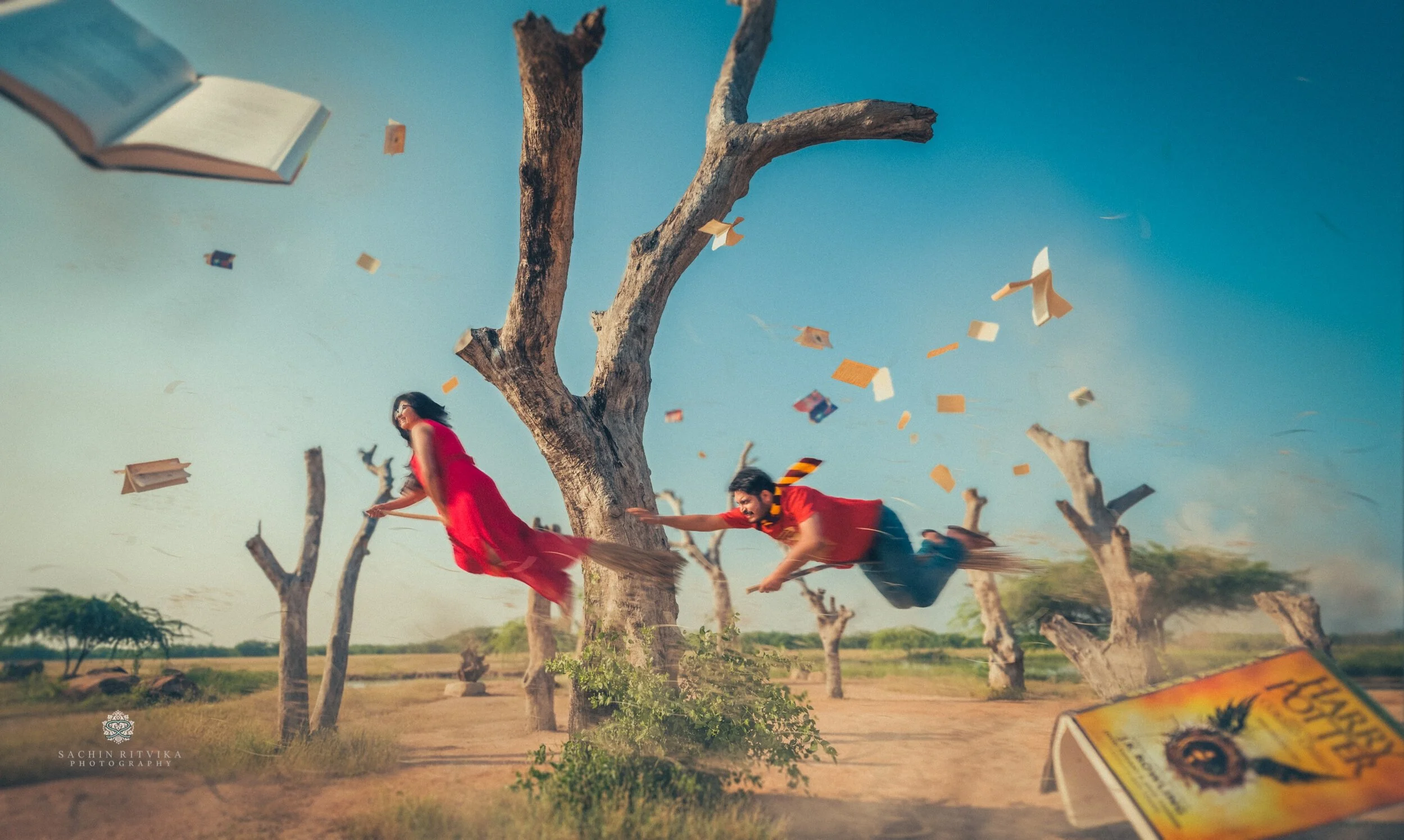Two people flying on broomsticks in a surreal scene with floating books and pages, set against a landscape with bare trees and a clear blue sky. One person wears a red dress, and the other wears a red shirt with a scarf. A "Harry Potter" book is visible in the foreground.