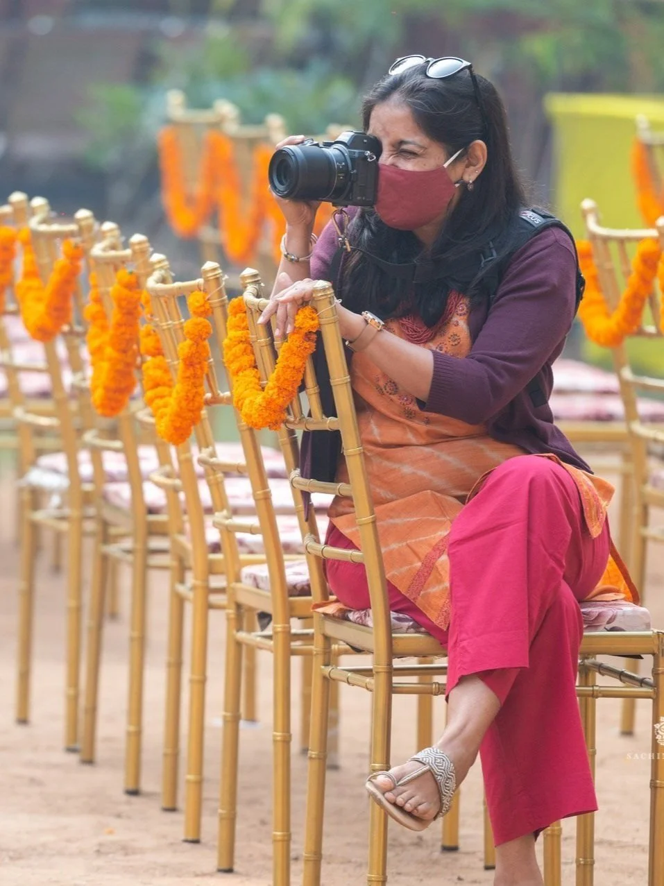 Ritvika shooting on gold-colored chairs decorated with marigold garlands, taking a photograph with a camera. She is wearing a face mask, sunglasses on her head, and dressed in colorful clothing. The setting appears to be an outdoor event.