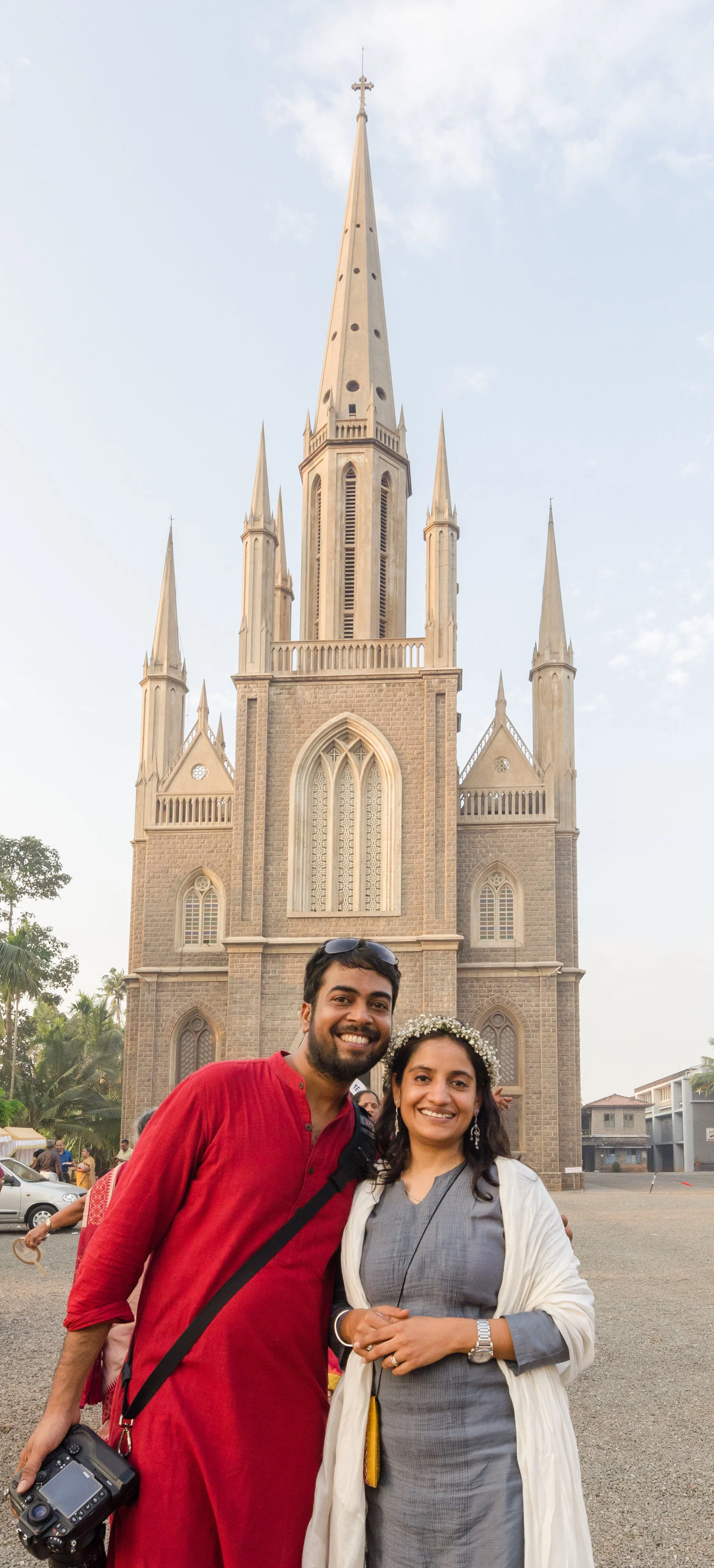 Two people smiling in front of a large, ornate church with tall spires and a pointed steeple, surrounded by trees and a clear sky.