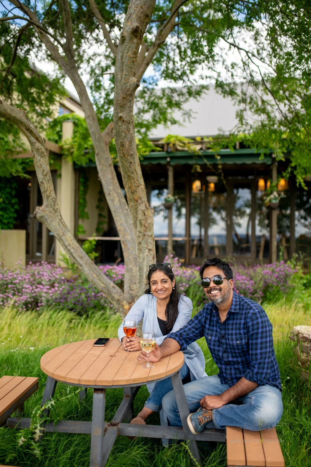 SachinRitvika sitting at a picnic table enjoying drinks, outdoors with greenery and a building in the background in Australian winery.