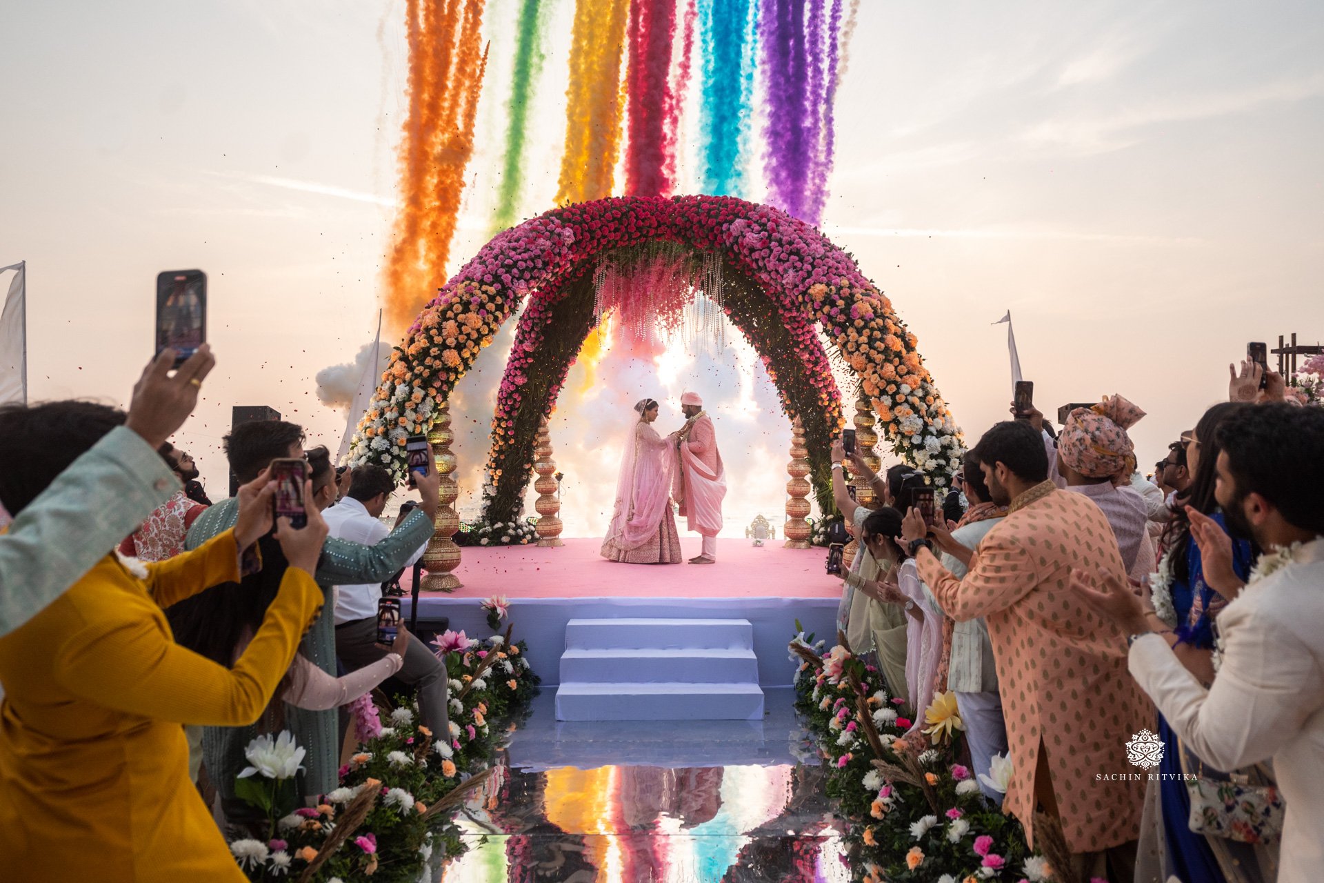 Wedding ceremony with floral arch and colorful smoke, bride and groom in traditional attire, guests taking photos.