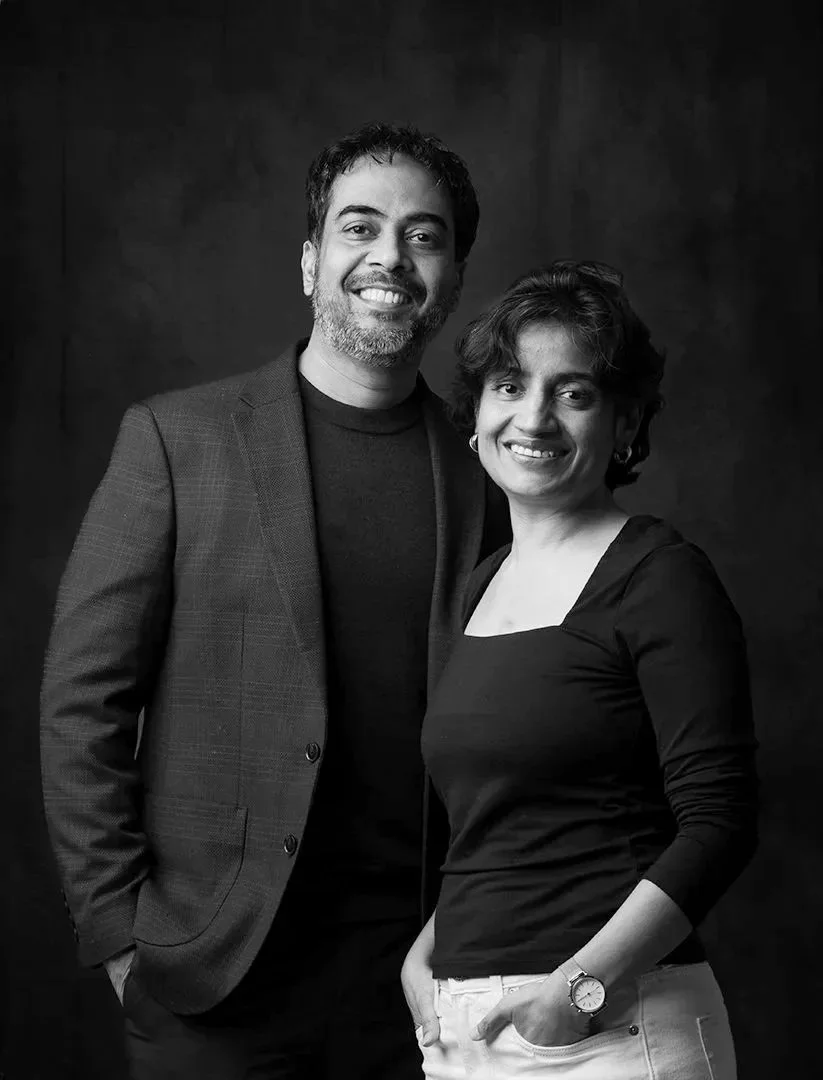 A black and white portrait of a man and a woman standing close together against a dark background. Both are smiling, with the man wearing a blazer over a dark shirt and the woman in a long-sleeved top and light-colored pants. The man has curly hair and facial hair, and the woman has short, wavy hair and is wearing a watch on her left wrist.