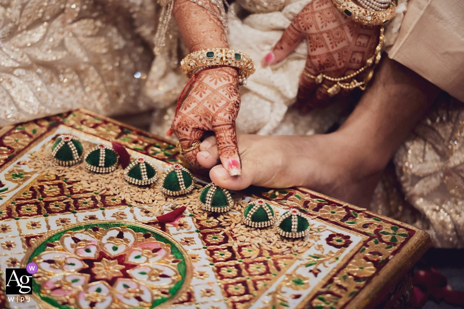 Close-up of a bride's hand with henna, adorned with jewelry, reaching for green and gold traditional earrings on an embroidered cloth, during an Indian wedding ceremony.