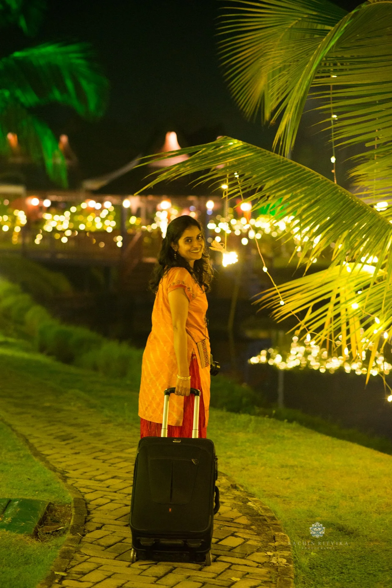 Ritvika in an orange dress pulling a suitcase on a garden path, surrounded by palm leaves and decorative lights at night.