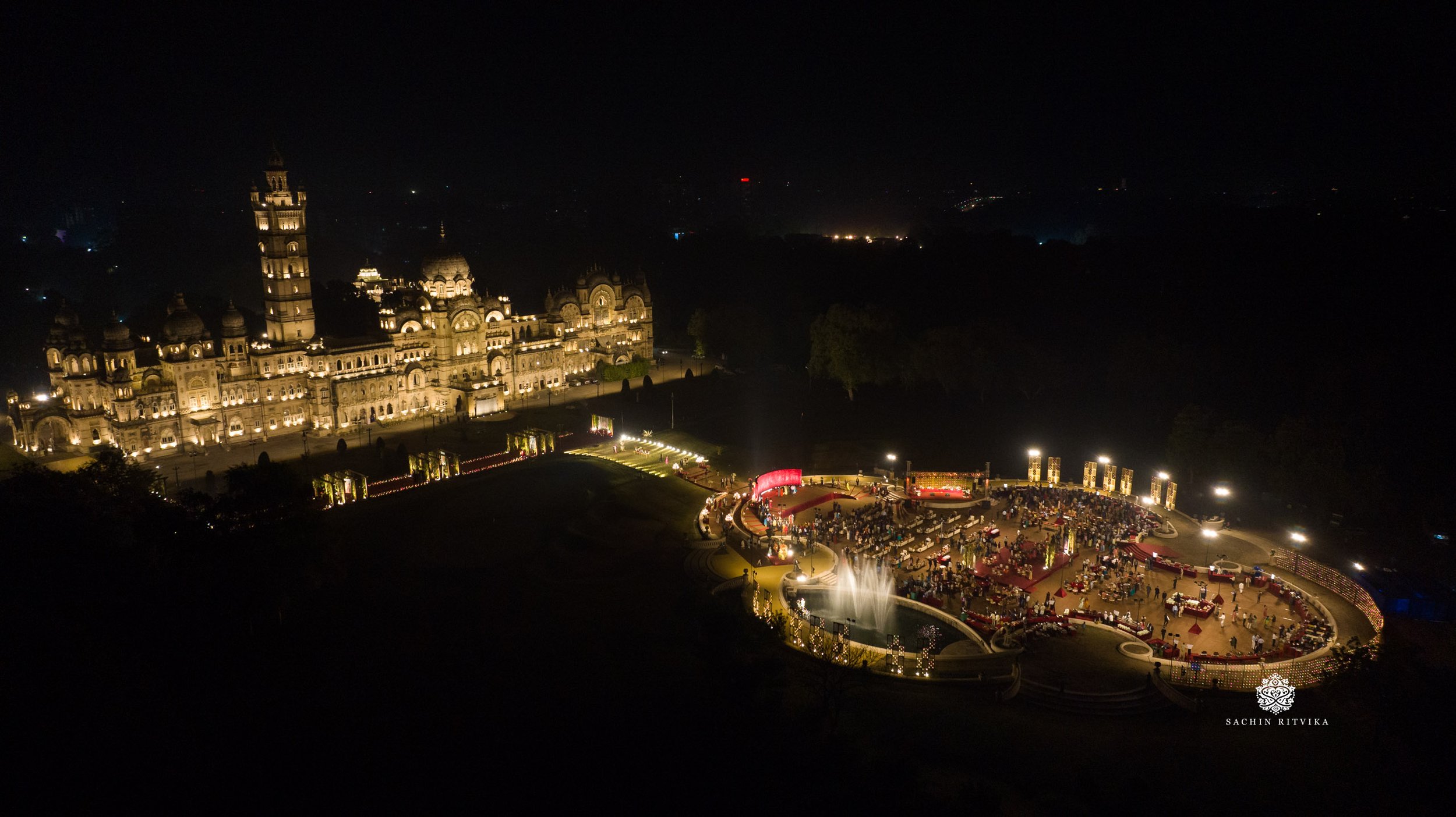 Aerial night view of palace illuminated with warm lighting. Next to the palace is a spacious, well-lit outdoor area with numerous people gathered, likely attending an event or celebration. The scene includes decorative lights and fountains, creating