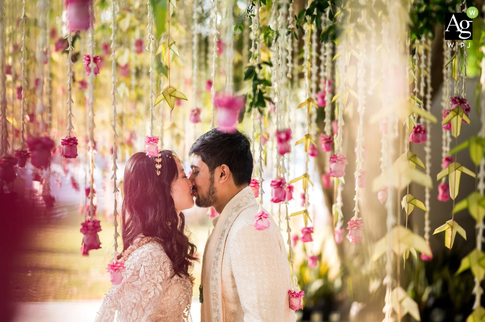 A couple in wedding attire sharing a kiss surrounded by hanging pink and white flower garlands.