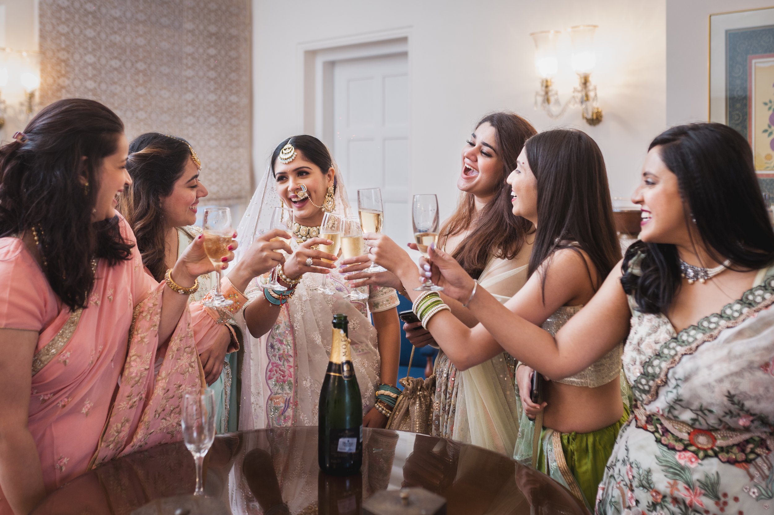 A group of women, with bride in centre in traditional attire toasting with champagne glasses indoors, celebrating joyfully around a table with a champagne bottle, at the heritage 7star property of Sukhvillas, Oberoi Hotels & Resorts, Chandigarh