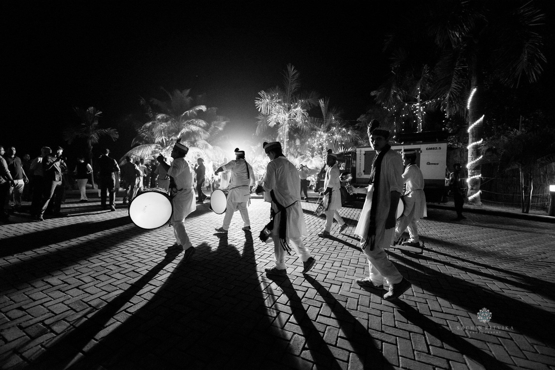A black and white photo of a traditional music procession with musicians playing drums, wearing traditional attire, and performing at an outdoor nighttime event with palm trees and lights in the background.