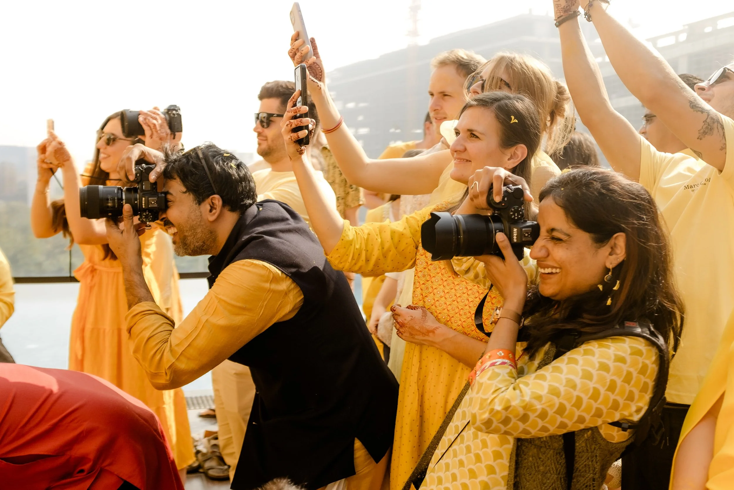 Photographers Sachinritvika smiling and joyfully capturing with a group of people in yellow clothing taking photos with cameras and smartphones at a haldi ceremony in india.