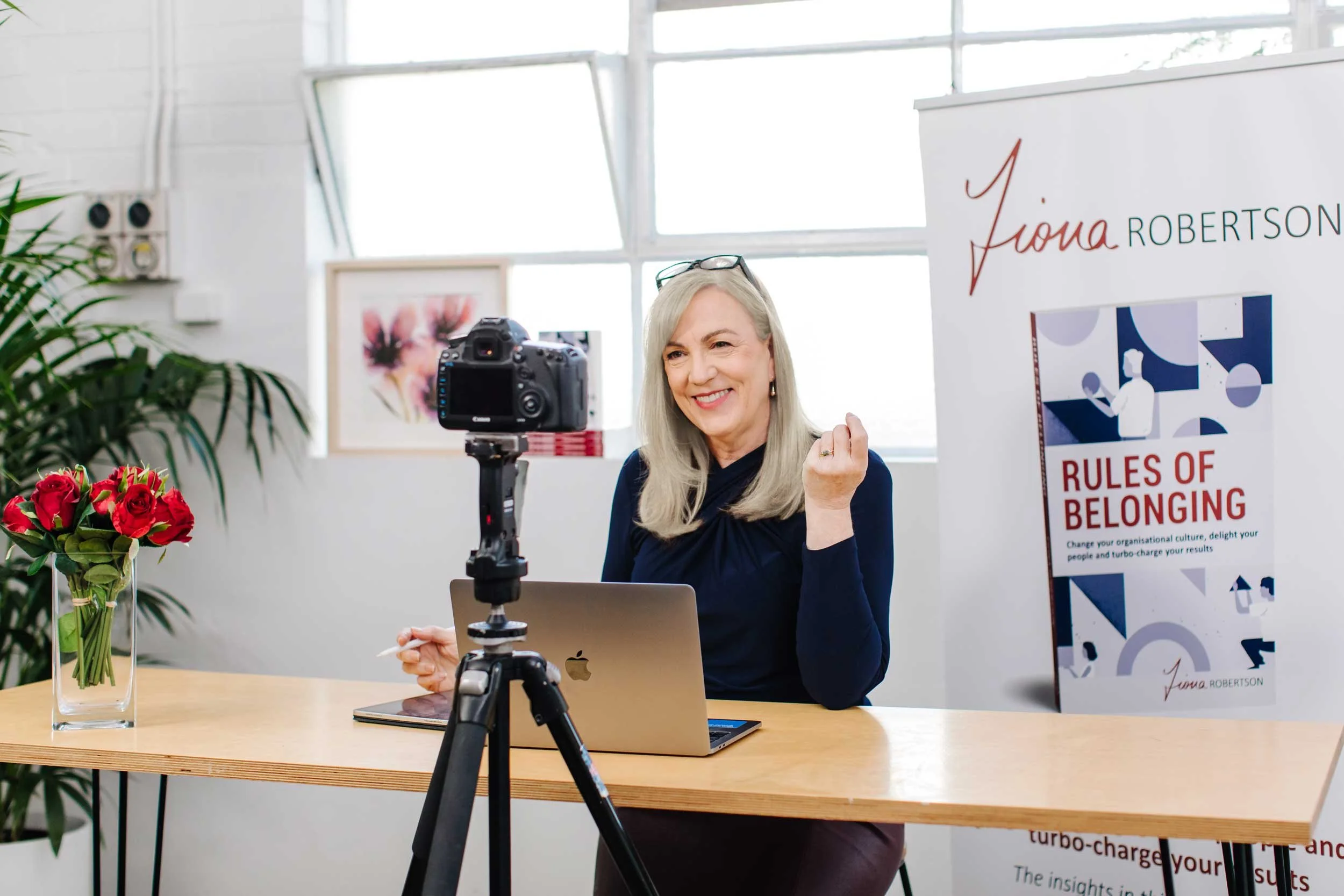 Fiona Robertson sitting at a table with a camera, features banner of Fiona's book Rules of Belonging.