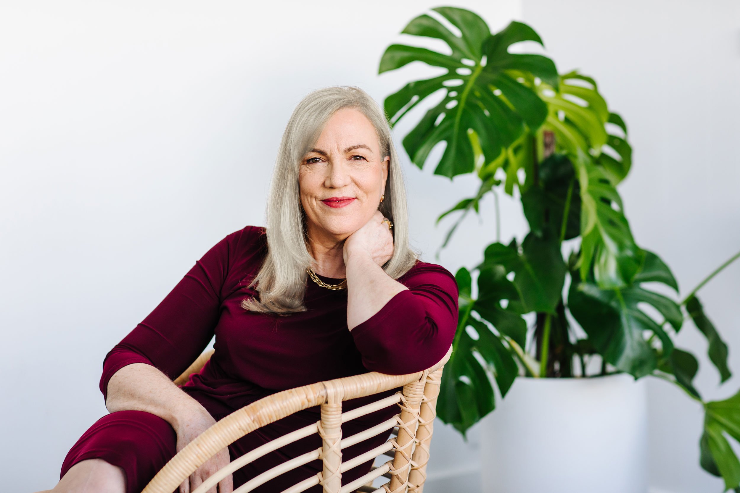 Fiona Robertson sitting in a cain chair indoors, smiling, surrounded by houseplants in a white pot. Fiona is wearing a warm red dress suitable for indoor relaxation.