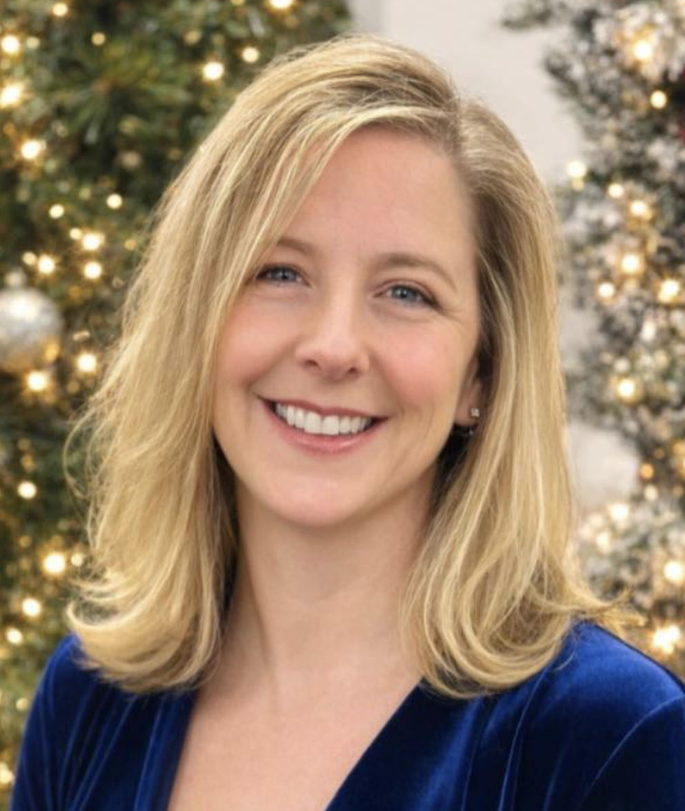 A smiling woman with shoulder-length blonde hair, wearing a blue top, stands in front of a decorated Christmas tree with lights.