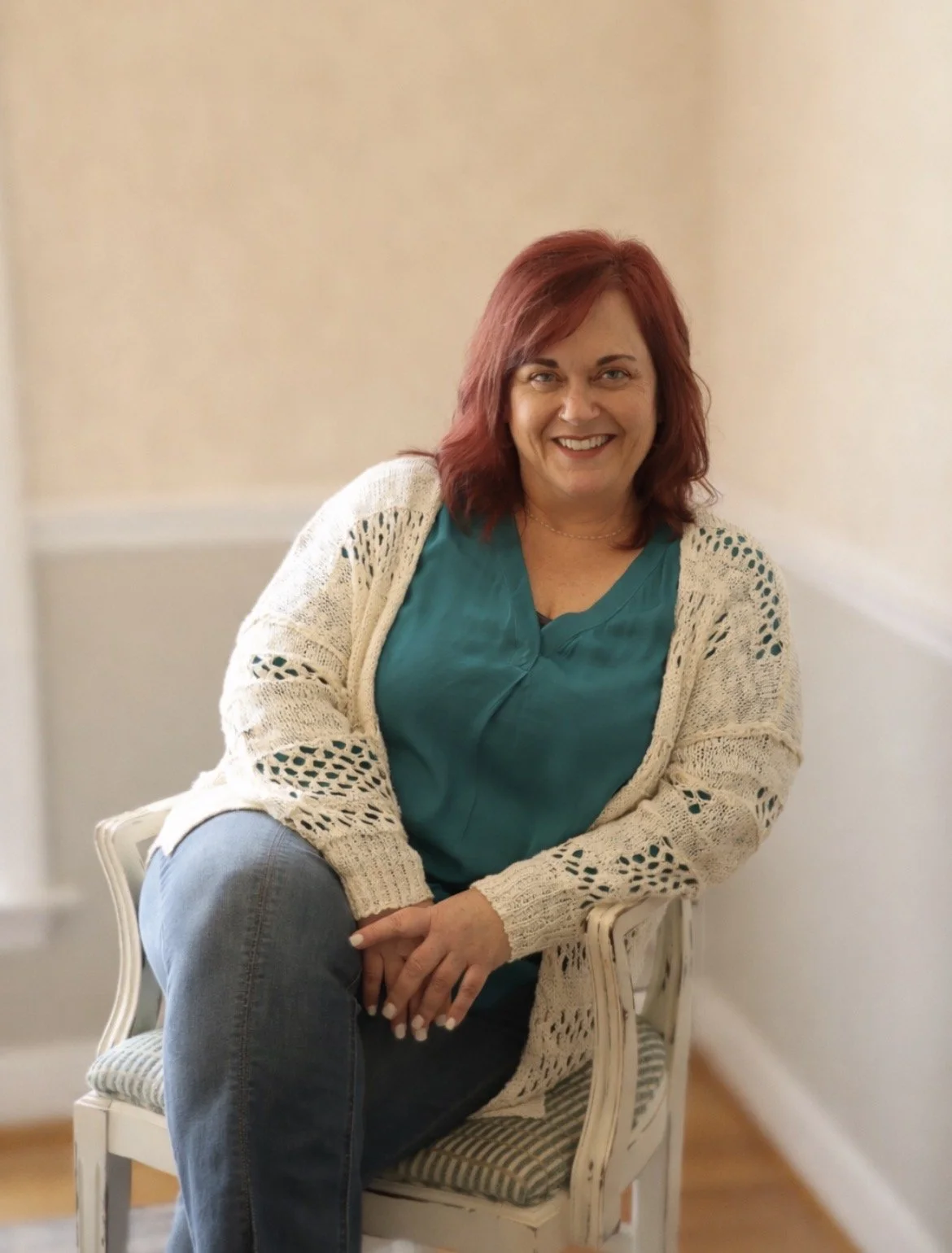 A woman with red hair smiling, sitting on a striped cushioned chair in a room with beige walls, wearing a green top, a cream open-knit cardigan, and jeans.