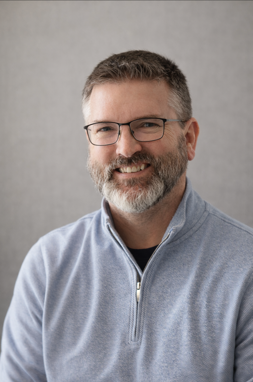 A middle-aged man with glasses, a beard, and short hair, smiling, wearing a light gray quarter-zip sweater against a plain gray background.