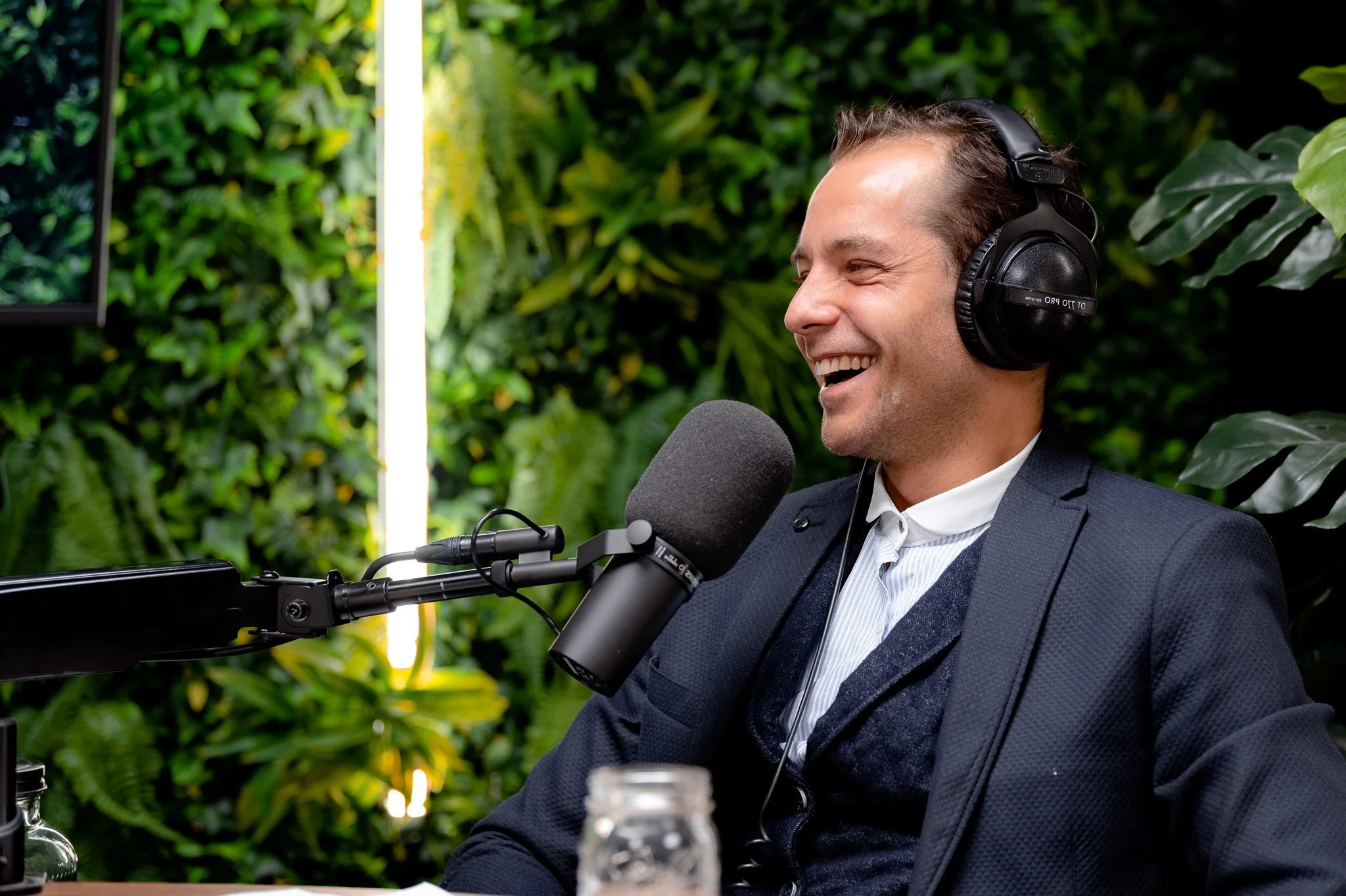 Man die een koptelefoon draagt en praat in een microfoon tijdens een opname, met een groene plantenwand op de achtergrond.