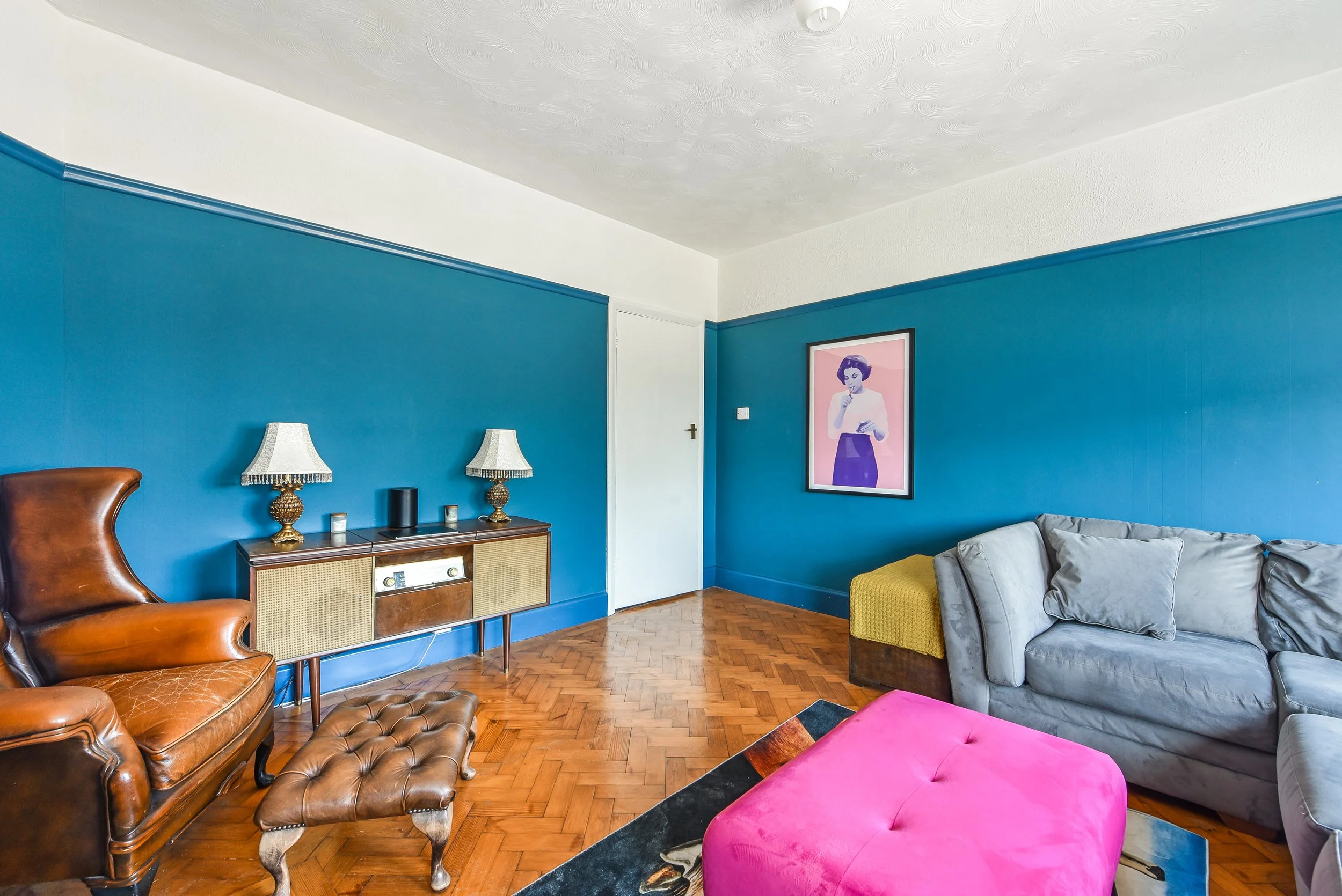 Living room with blue walls, hardwood floor, brown leather armchair and matching footstool, gray couch, colorful pink ottoman, framed art of a woman, and a wooden sideboard with lamps and decorative items.