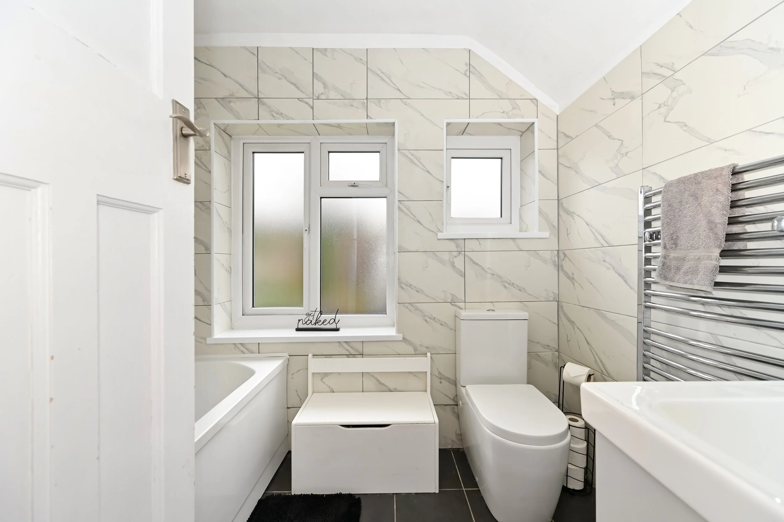 Bathroom with white tiled walls, a bathtub below three windows, a toilet, a towel heated rack with a gray towel, and a window seat with a black mat.