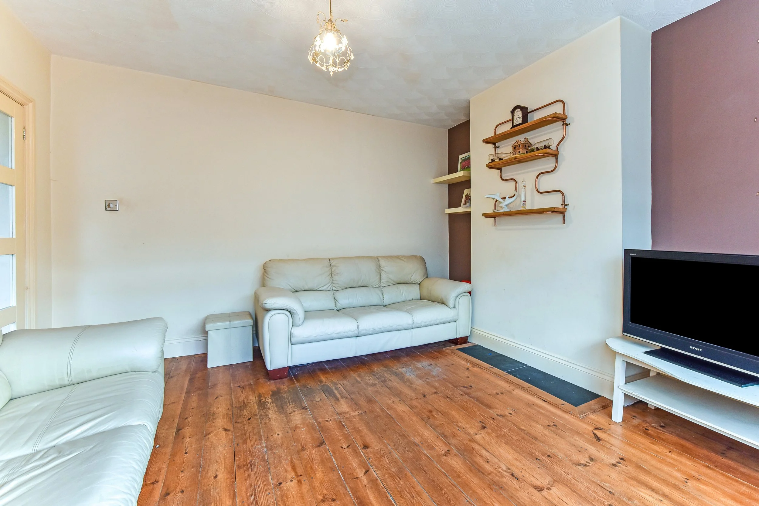 Living room with wooden floors, two white leather sofas, a wall-mounted shelving unit with decorative items, a small TV on a white stand, and a fire vent near the floor.