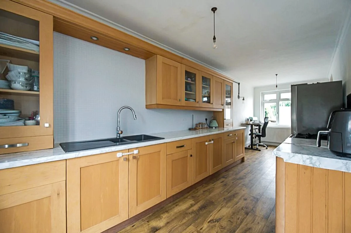 Kitchen with wooden cabinets, marble countertops, a black sink, and various appliances, including a refrigerator, toaster, and kettle, with a window at the end of the room.