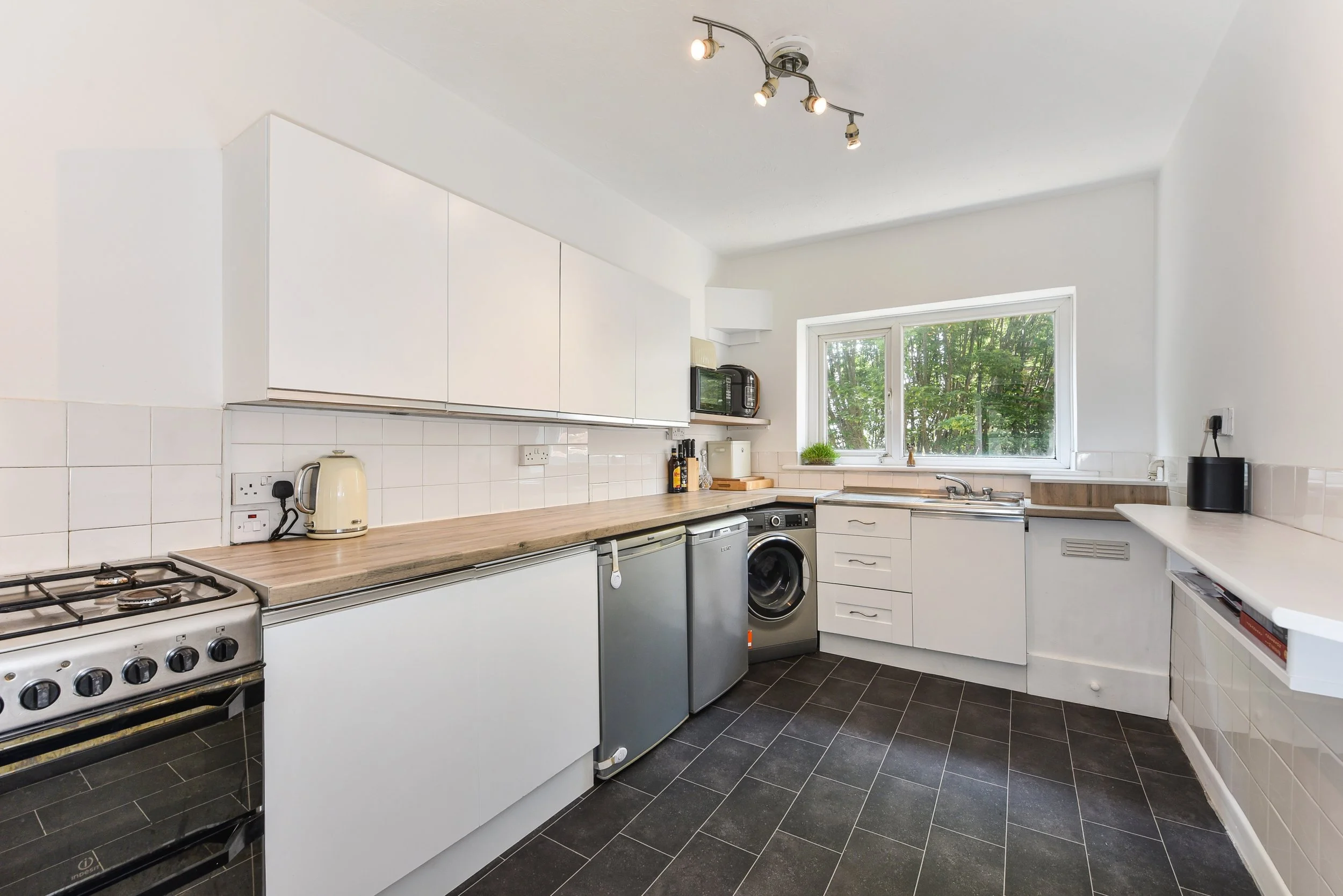 Kitchen with white cabinets, black tile floor, window overlooking greenery, appliances including stove, microwave, toaster, washing machine, and small refrigerator.