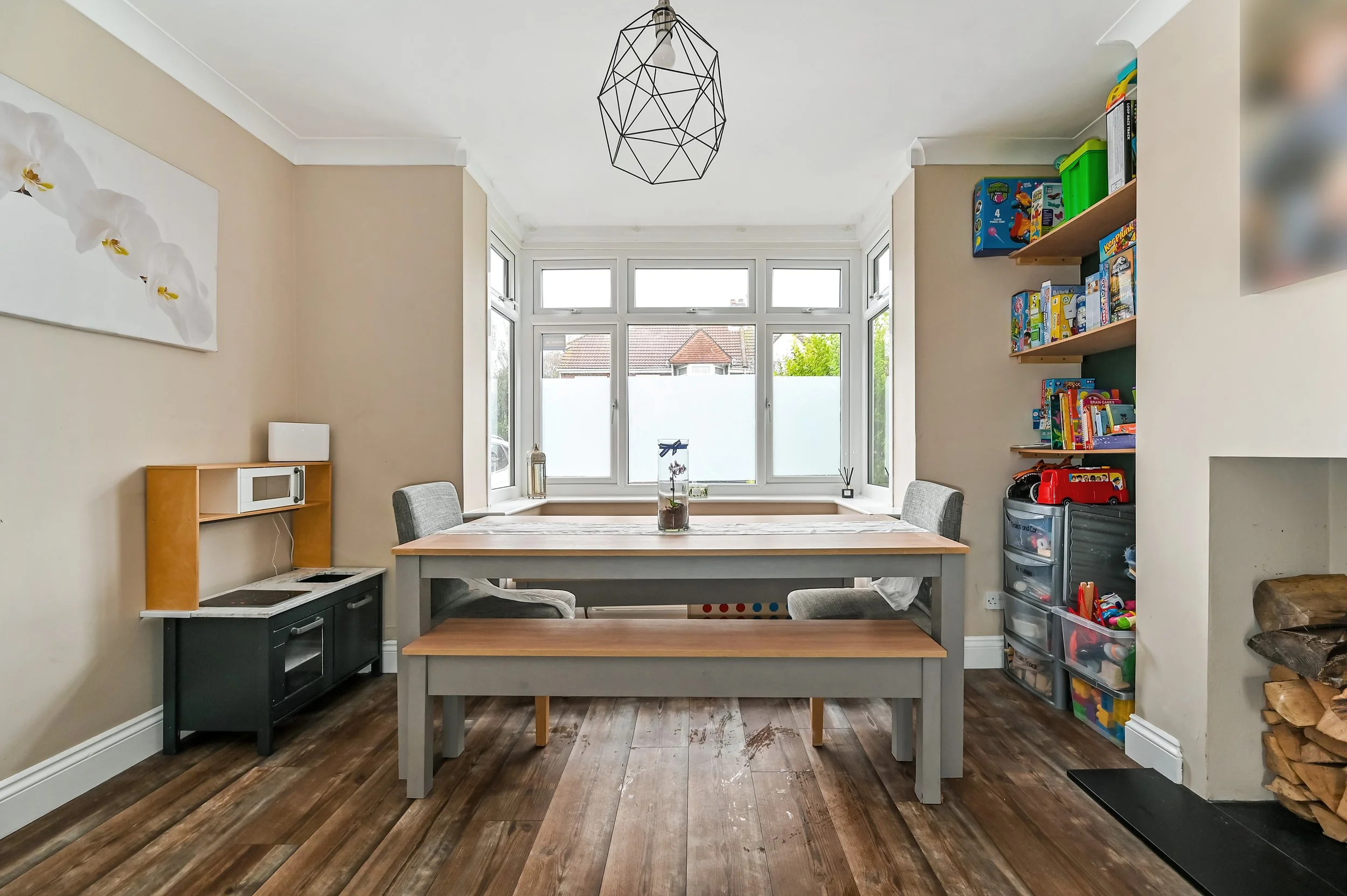 Dining area with a wooden table and bench, two chairs, a corner shelf with a microwave, and a colorful toy organizer near a fireplace with logs. Large windows with frosted glass.