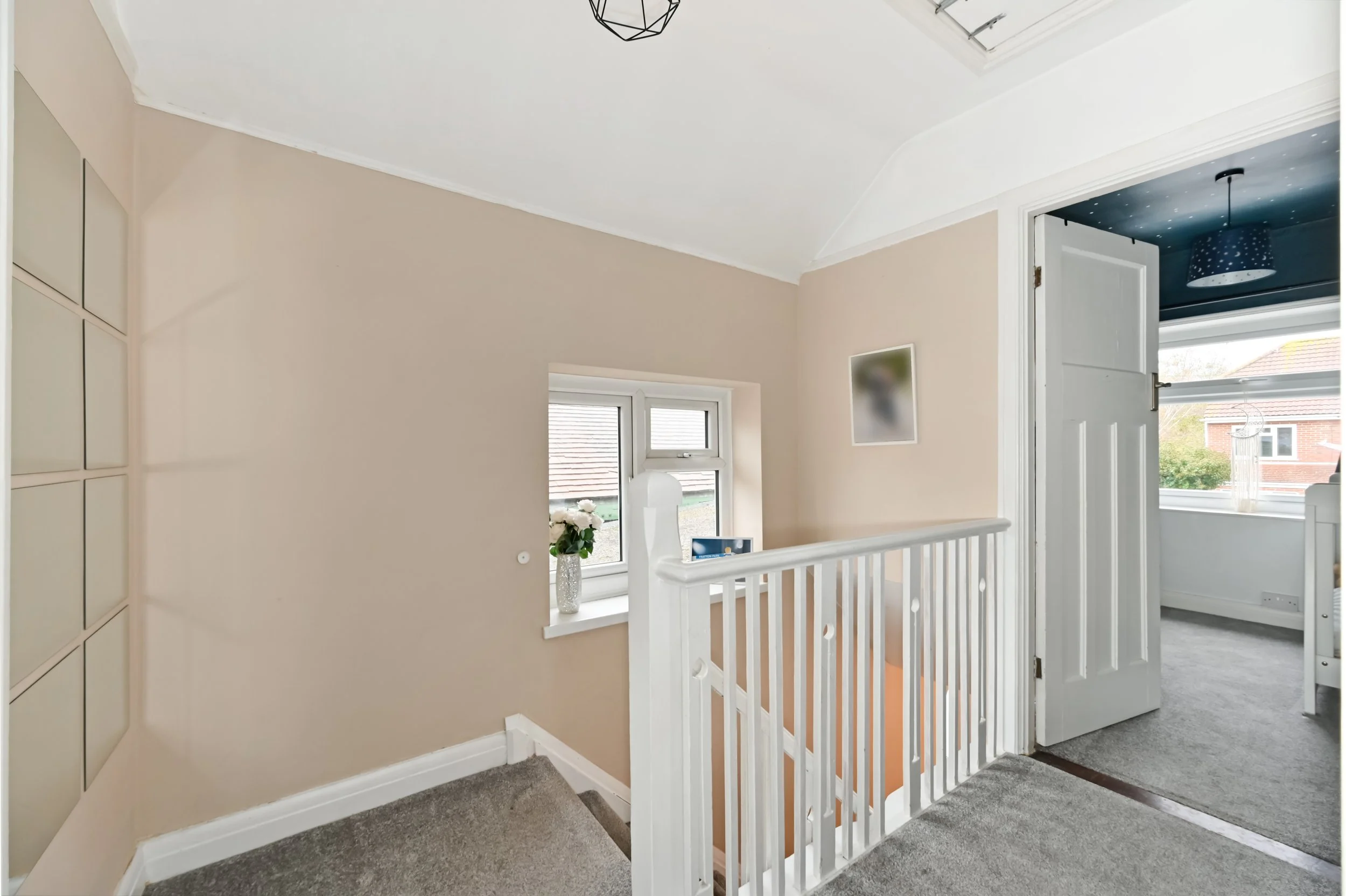 An interior view of a upper floor landing area with a staircase, beige walls, a small window with a plant on the windowsill, and an open door leading to a room with a dark blue ceiling and a window.