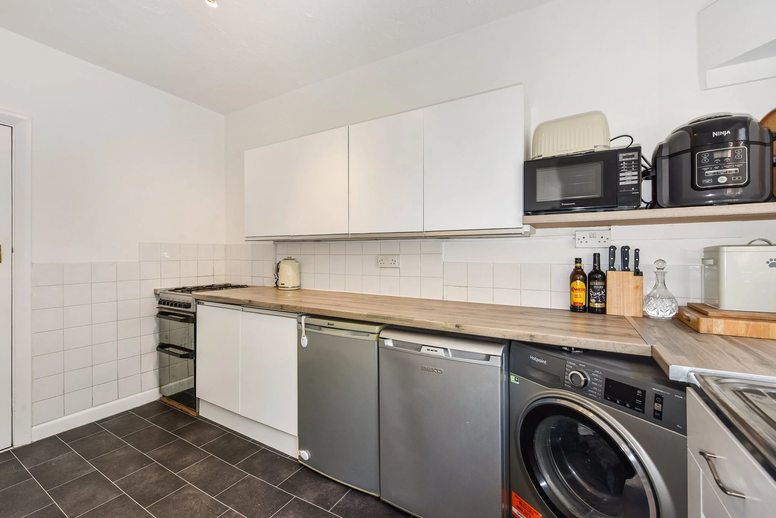 Kitchen with white cabinets, black oven, mini fridge, washing machine, microwave, rice cooker, and various kitchen items on the counter.