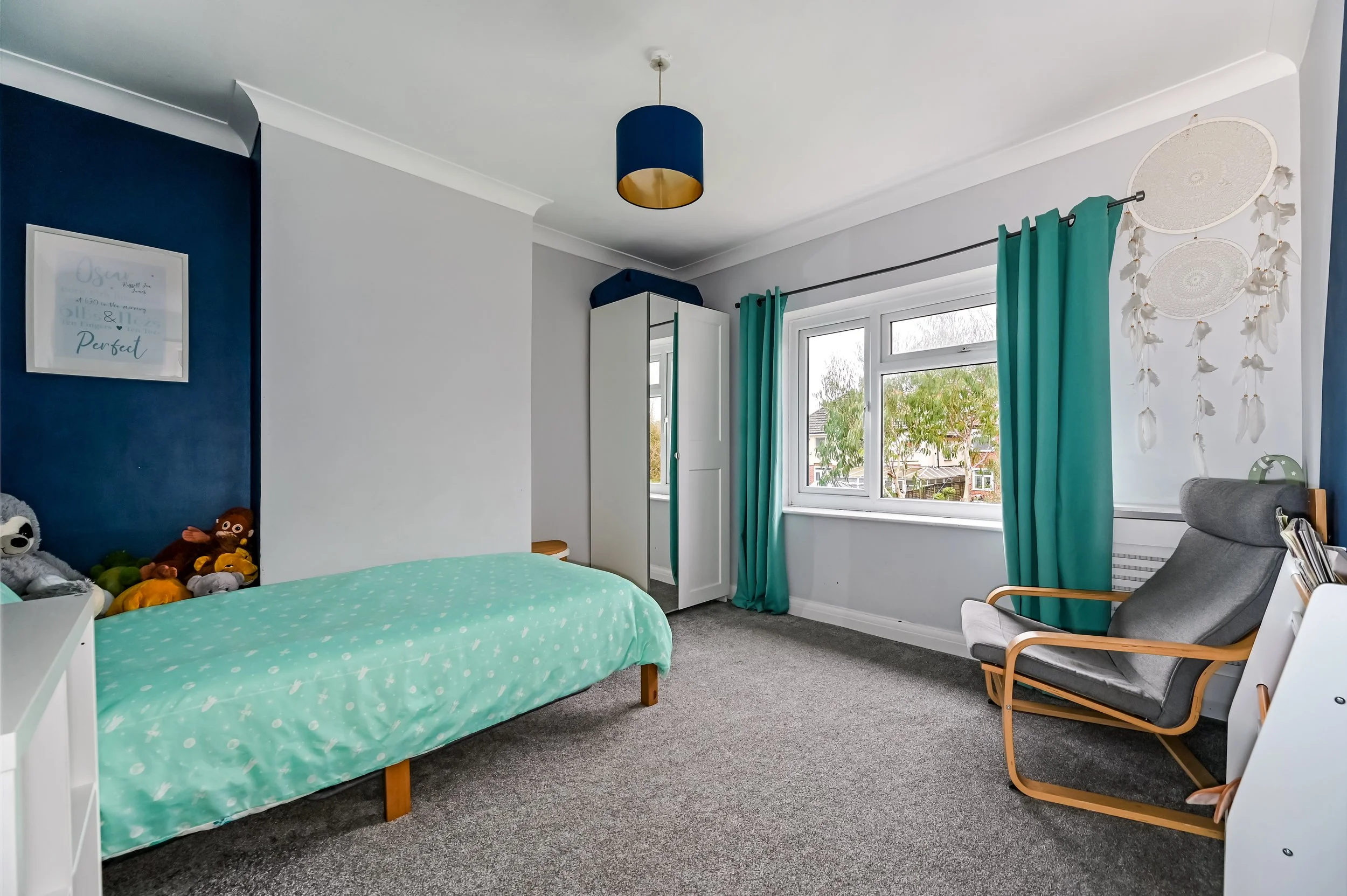 A bedroom with a single bed, toys, a white wardrobe, a window with green curtains, a gray chair, a decorative wall hanging, and framed artwork.