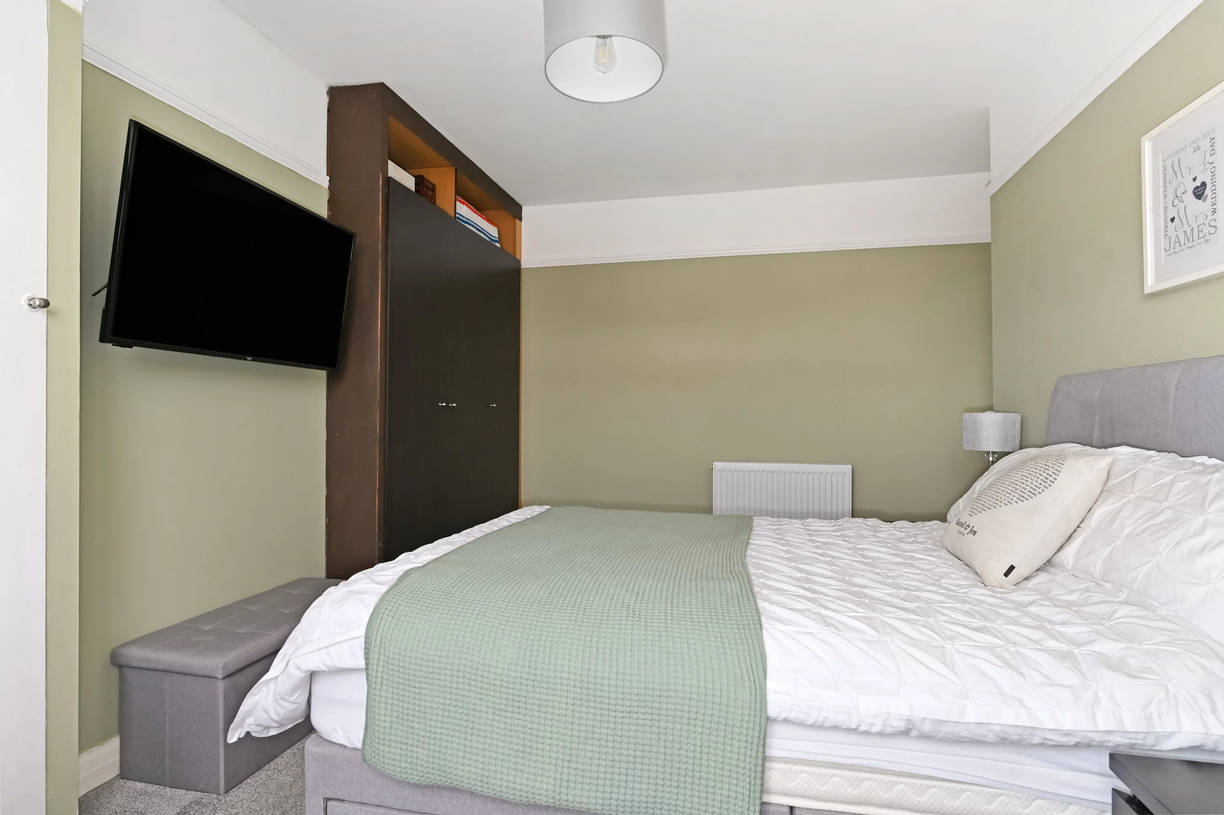 A bedroom with a bed, green blanket, gray headboard, cushion, wall-mounted TV on the left, small gray bench at the foot of the bed, nightstand with a lamp, and framed artwork on the wall