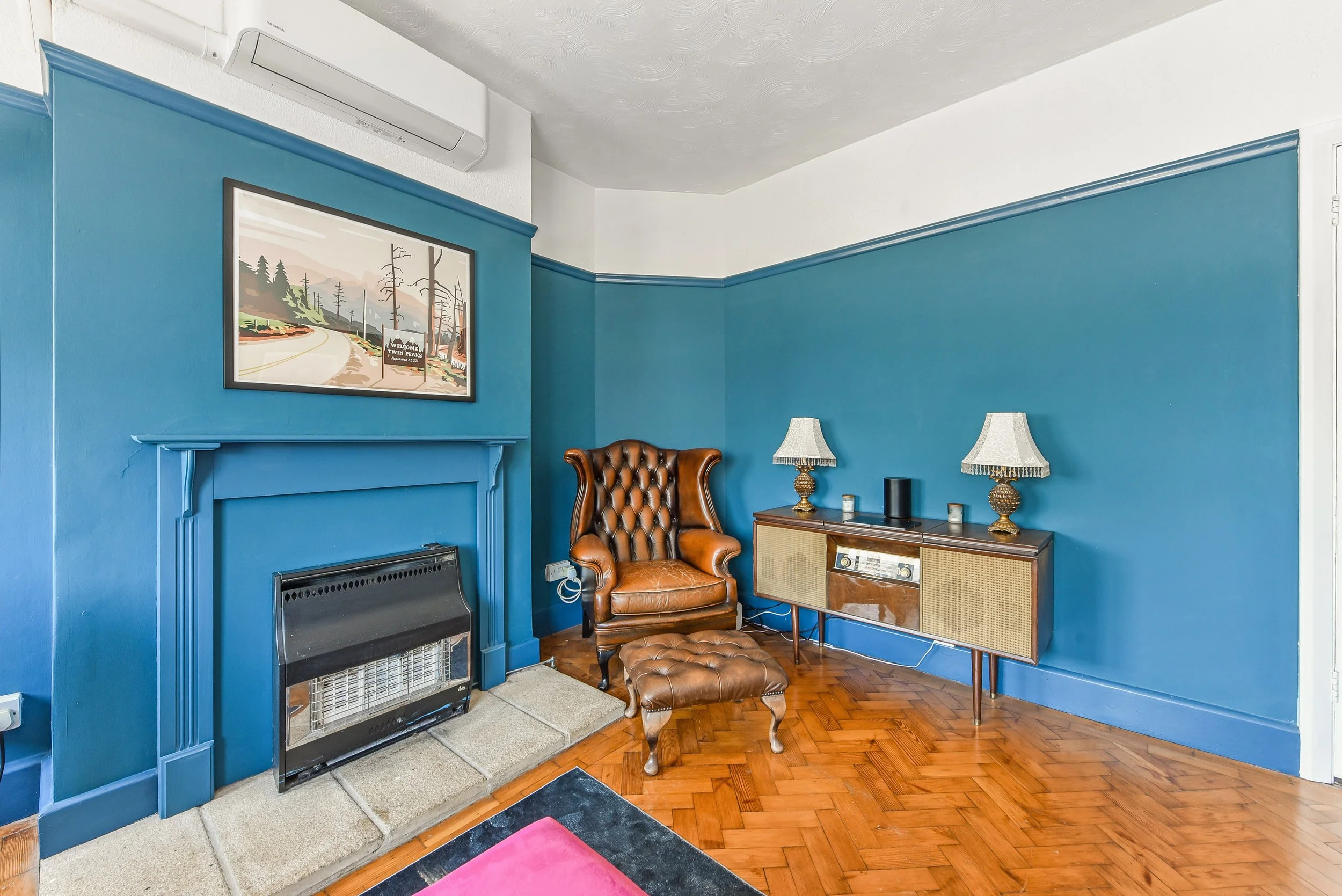 Living room with blue walls, a fireplace with a black heater, a brown leather armchair with matching ottoman, a vintage wooden sideboard with two lamps, and a framed landscape painting on the wall.