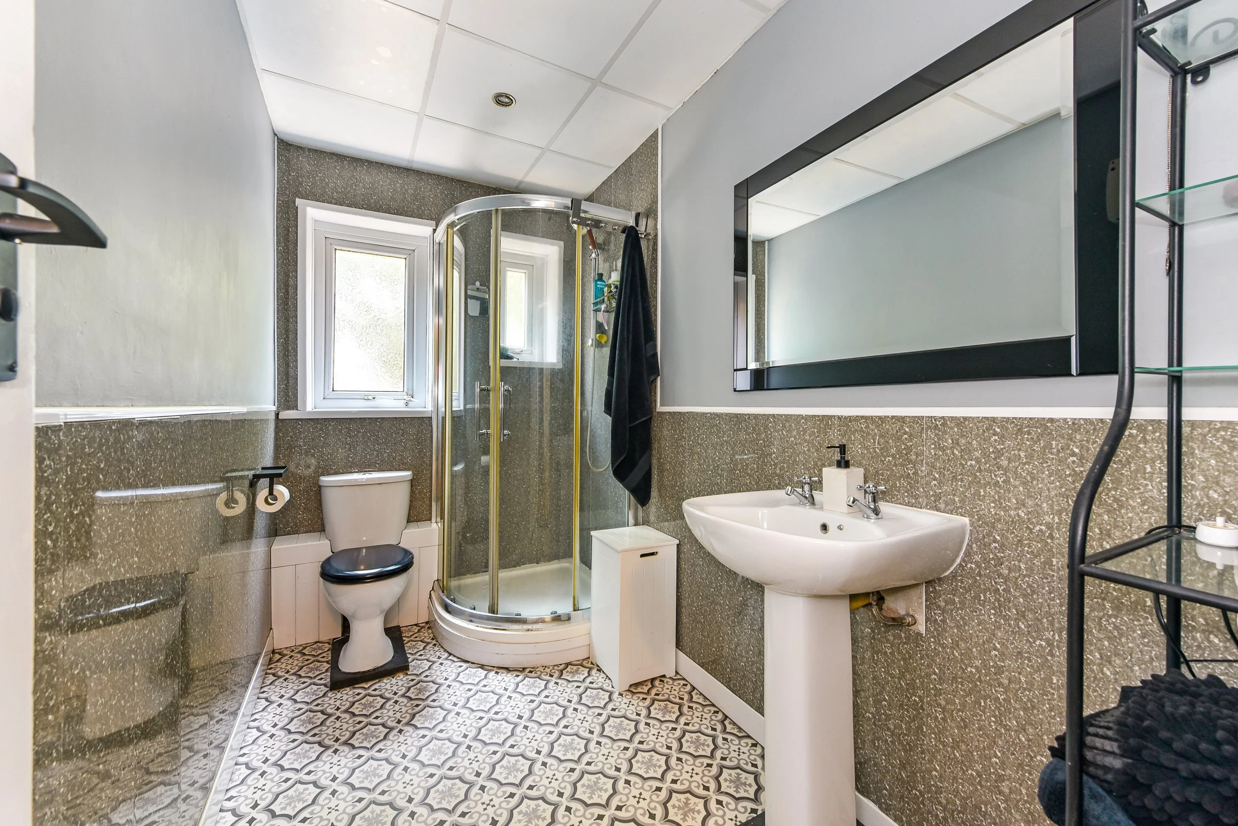 Bathroom with small shower enclosure, toilet, sink, large mirror, and decorative floor tiles.