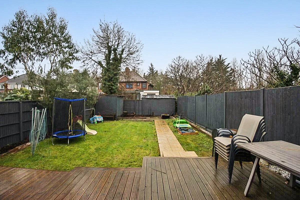Backyard with wooden deck, stack of chairs, plastic table, and outdoor toys including a trampoline, slide, and sandbox, enclosed by a dark fence, with trees and neighboring houses in the background under a clear sky.