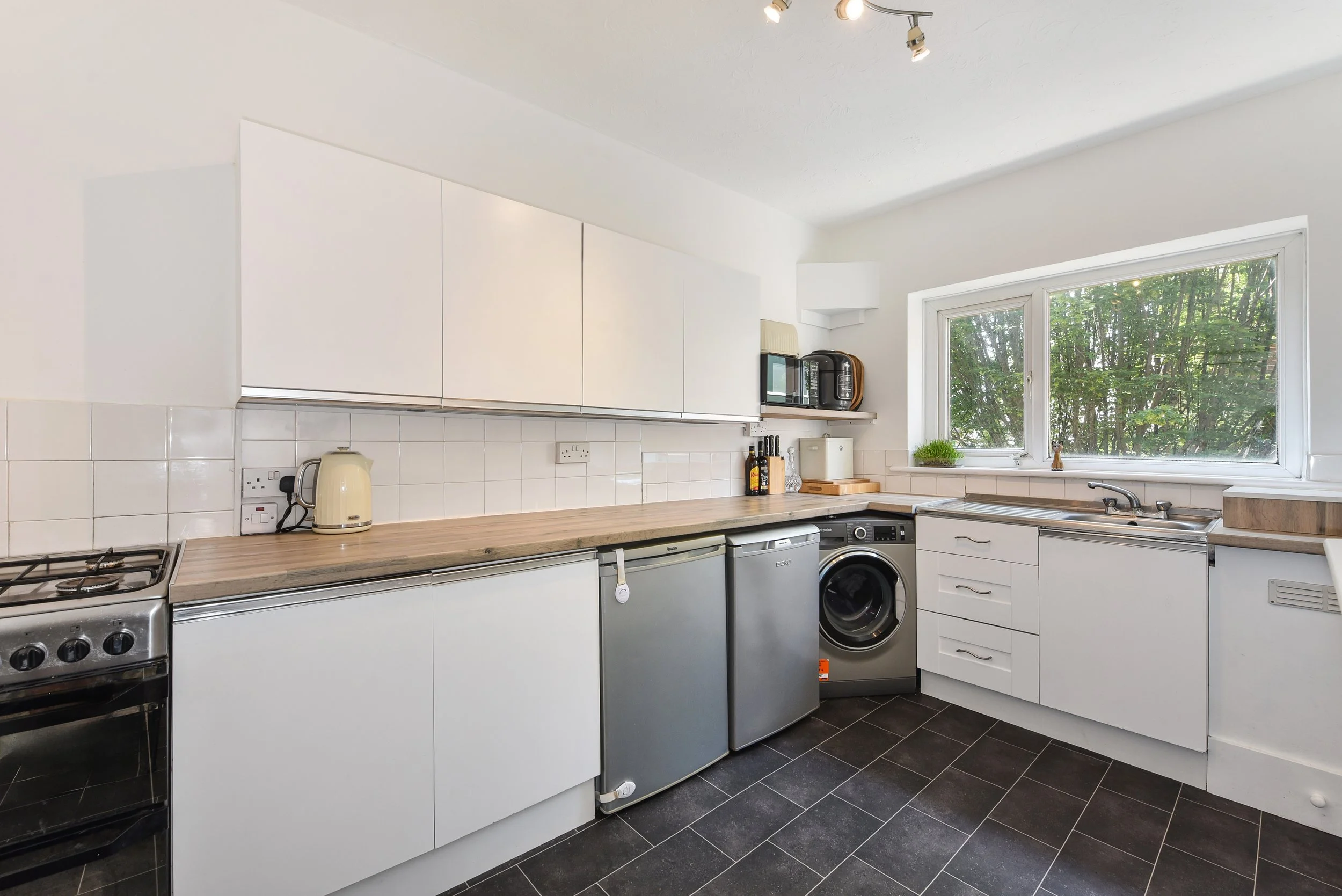 Kitchen with white cabinets, black floor tiles, a window above the sink, and appliances including a stove, mini fridge, washing machine, microwave, and kettle.