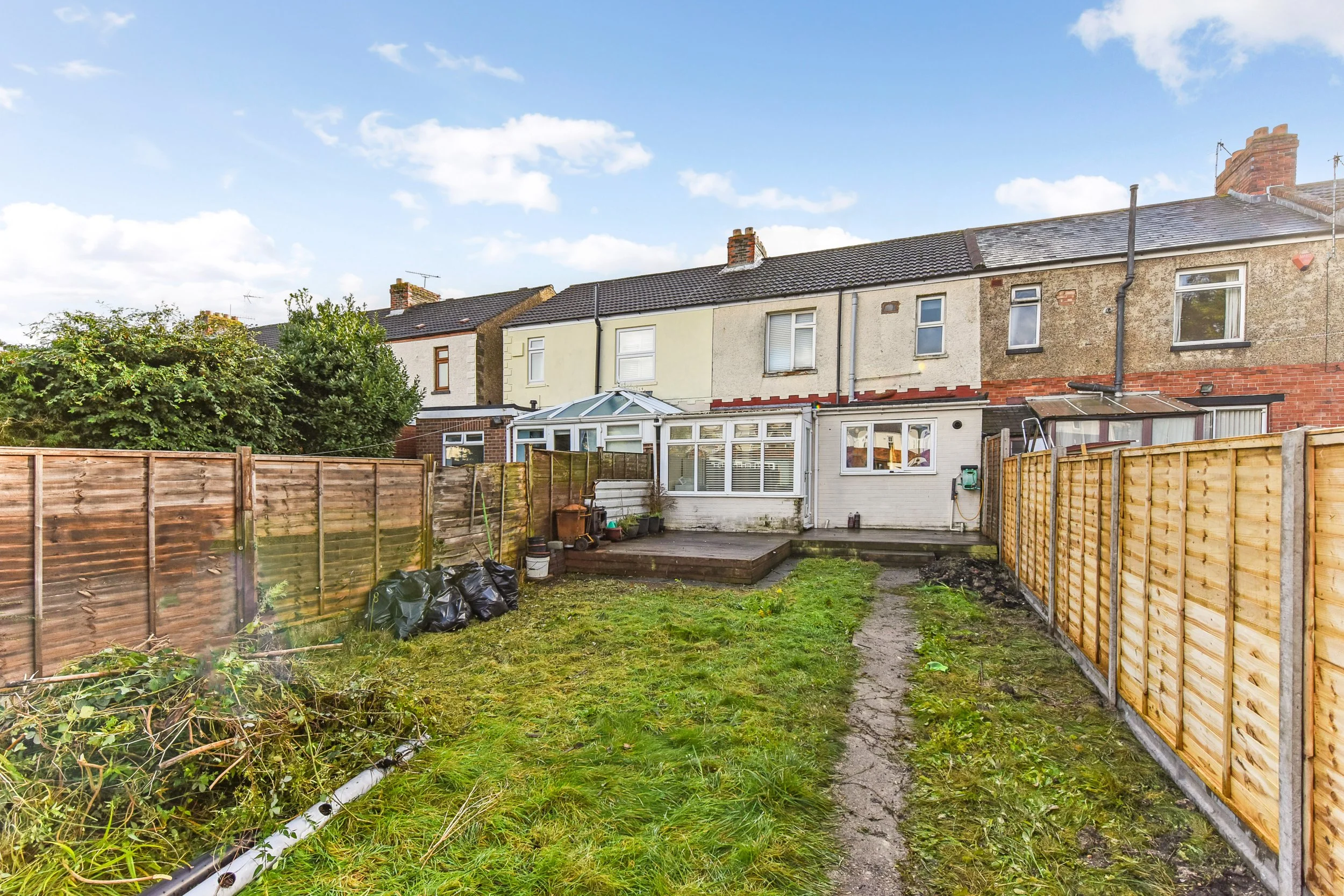 Backyard with grass and a narrow dirt path, surrounded by wooden fences, with a row of terraced houses with brick and stucco exteriors and a greenhouse on the back porch.