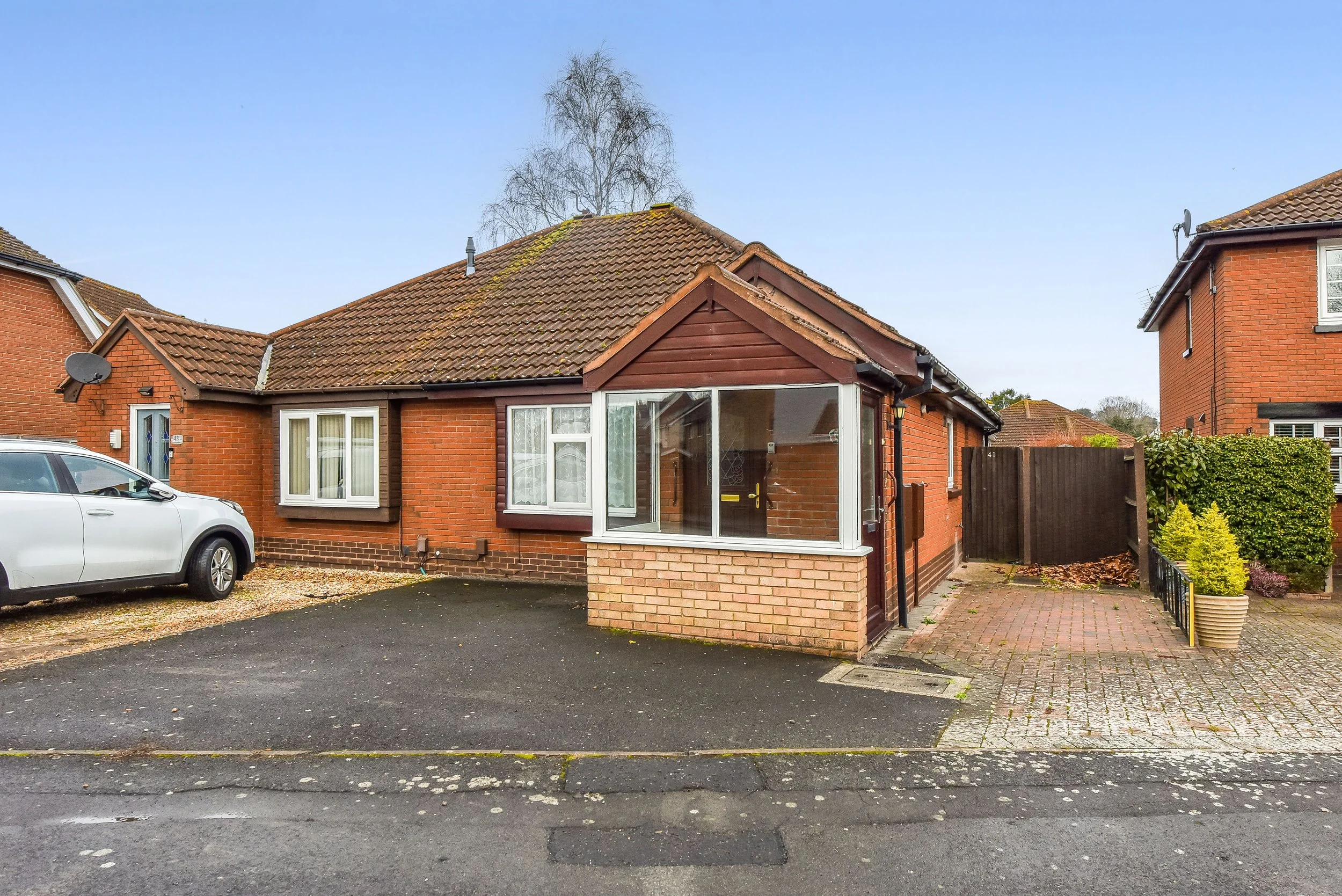 A single-story brick house with a brown tiled roof, a small enclosed front porch with large windows, a driveway with a white car parked, and a pathway with garden plants to the side.